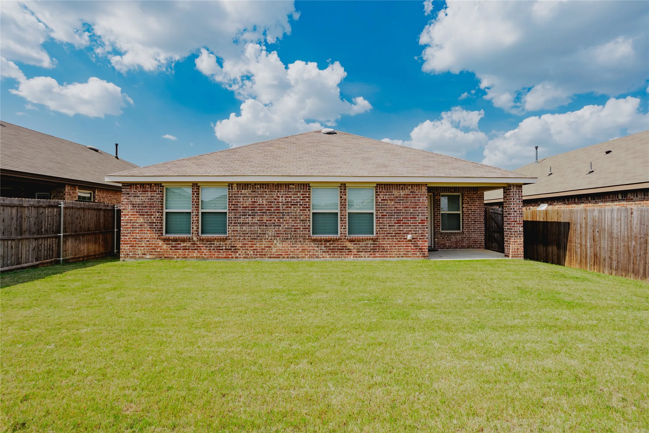 Expansive backyard featuring a manicured lawn, full brick exterior, multiple windows with white trim, a covered patio, and a privacy fence
