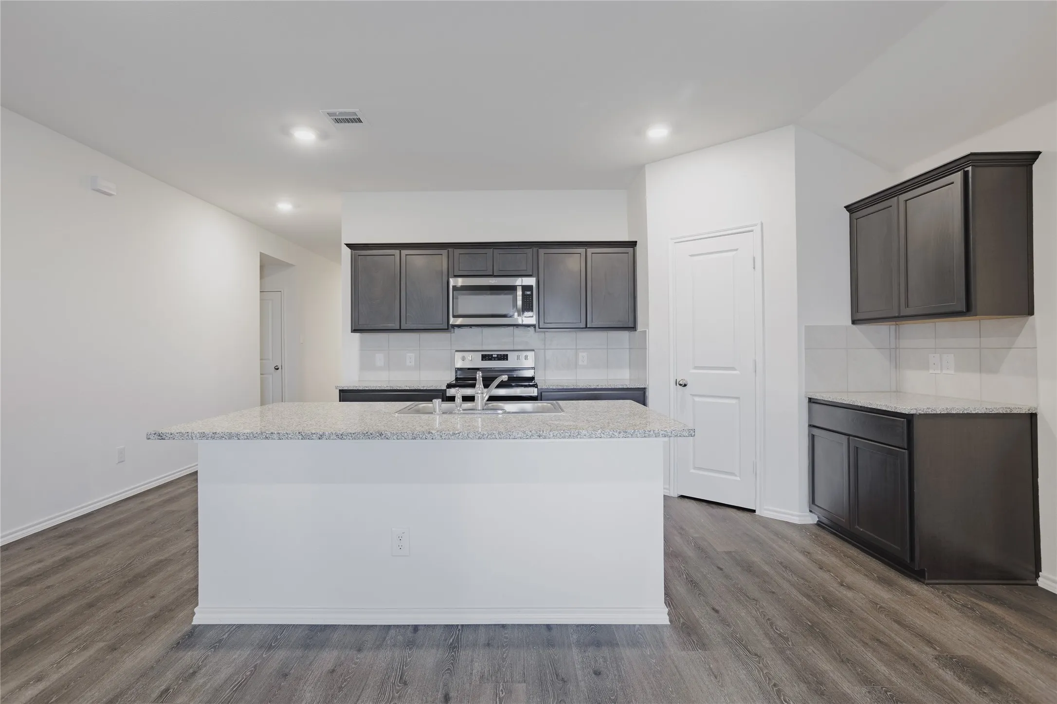 Open-concept kitchen featuring an island with a light-toned countertop, dark wood-finish cabinetry, stainless steel appliances, a white tile backsplash, and wood-finish flooring