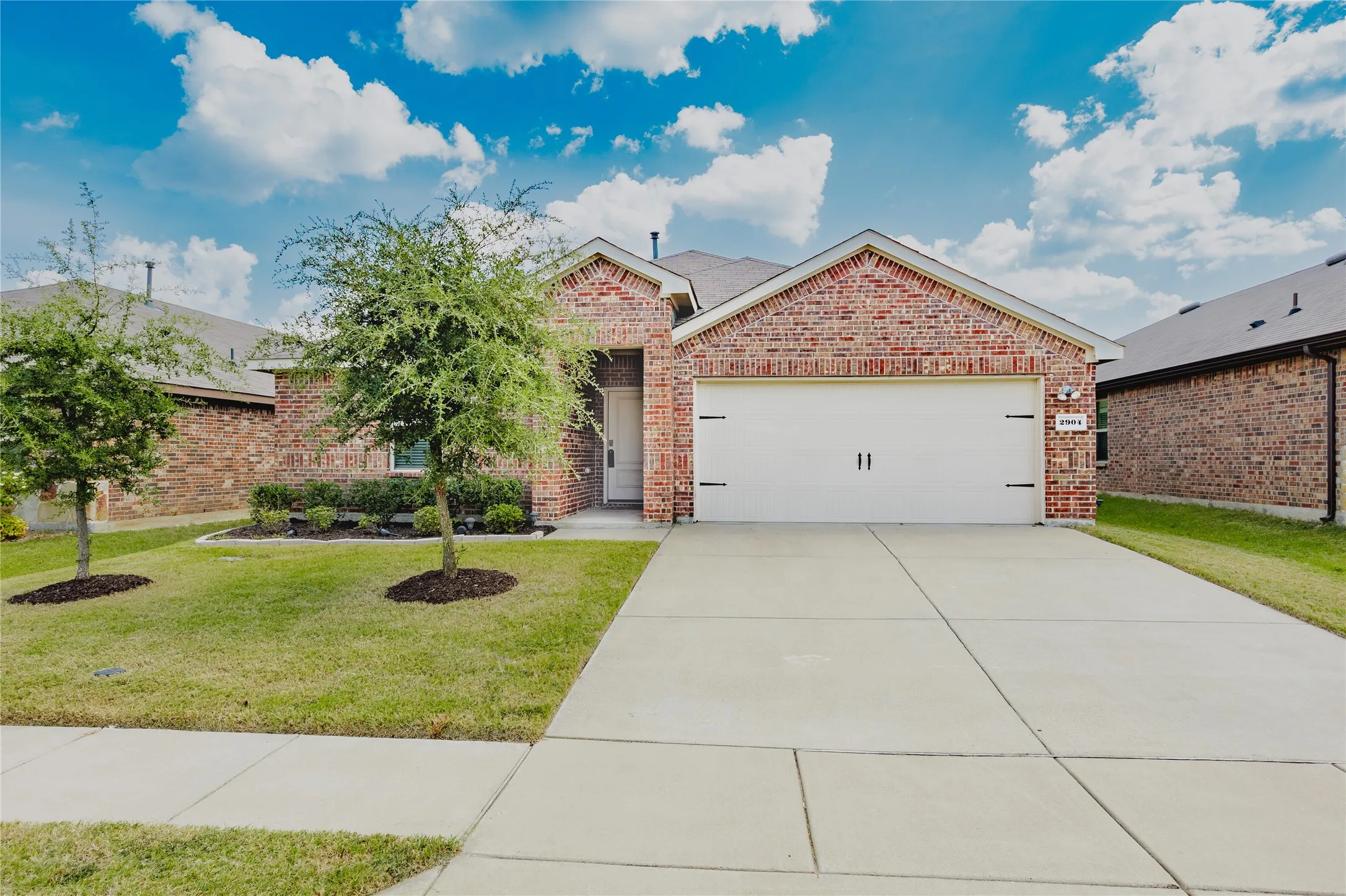 Brick exterior featuring a two-car garage with decorative hardware, an extended concrete driveway, and a manicured lawn with mature trees