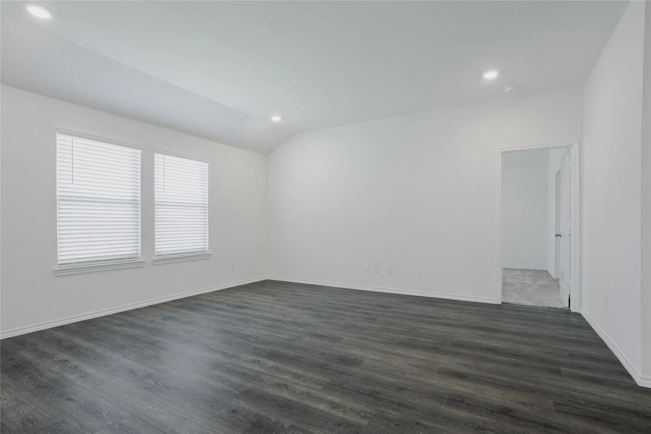 Spacious room featuring dark wood-finish flooring, two windows with blinds, recessed lighting, and white walls