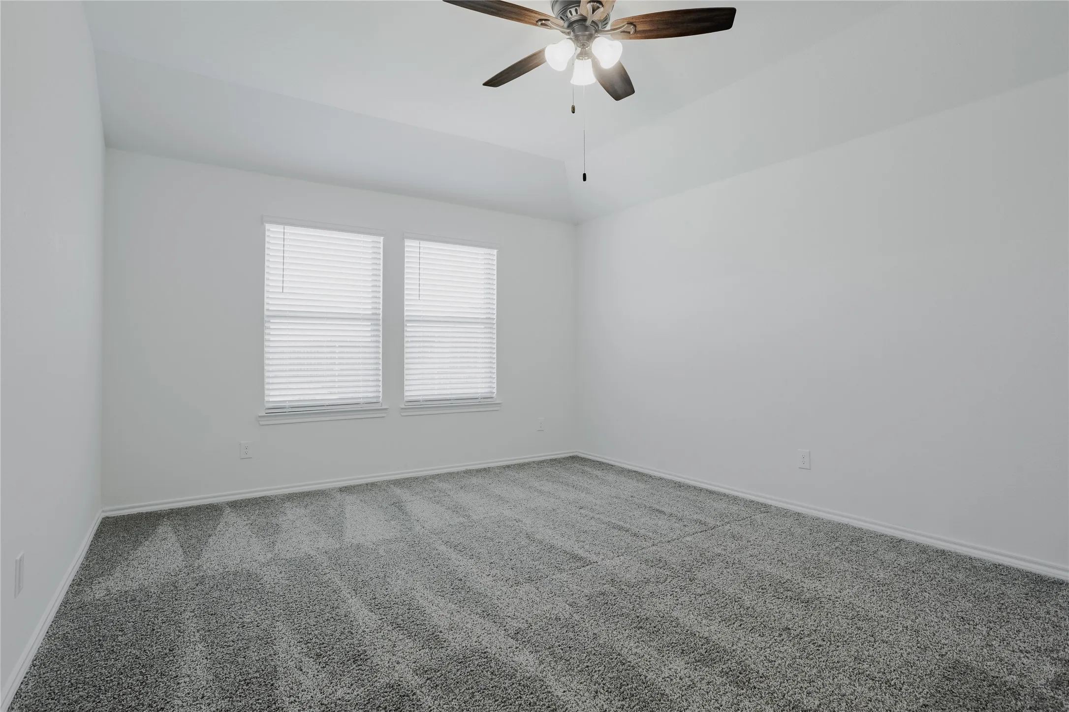 Spacious room featuring neutral carpeting, a ceiling fan with integrated lighting, two windows with blinds, and white painted walls