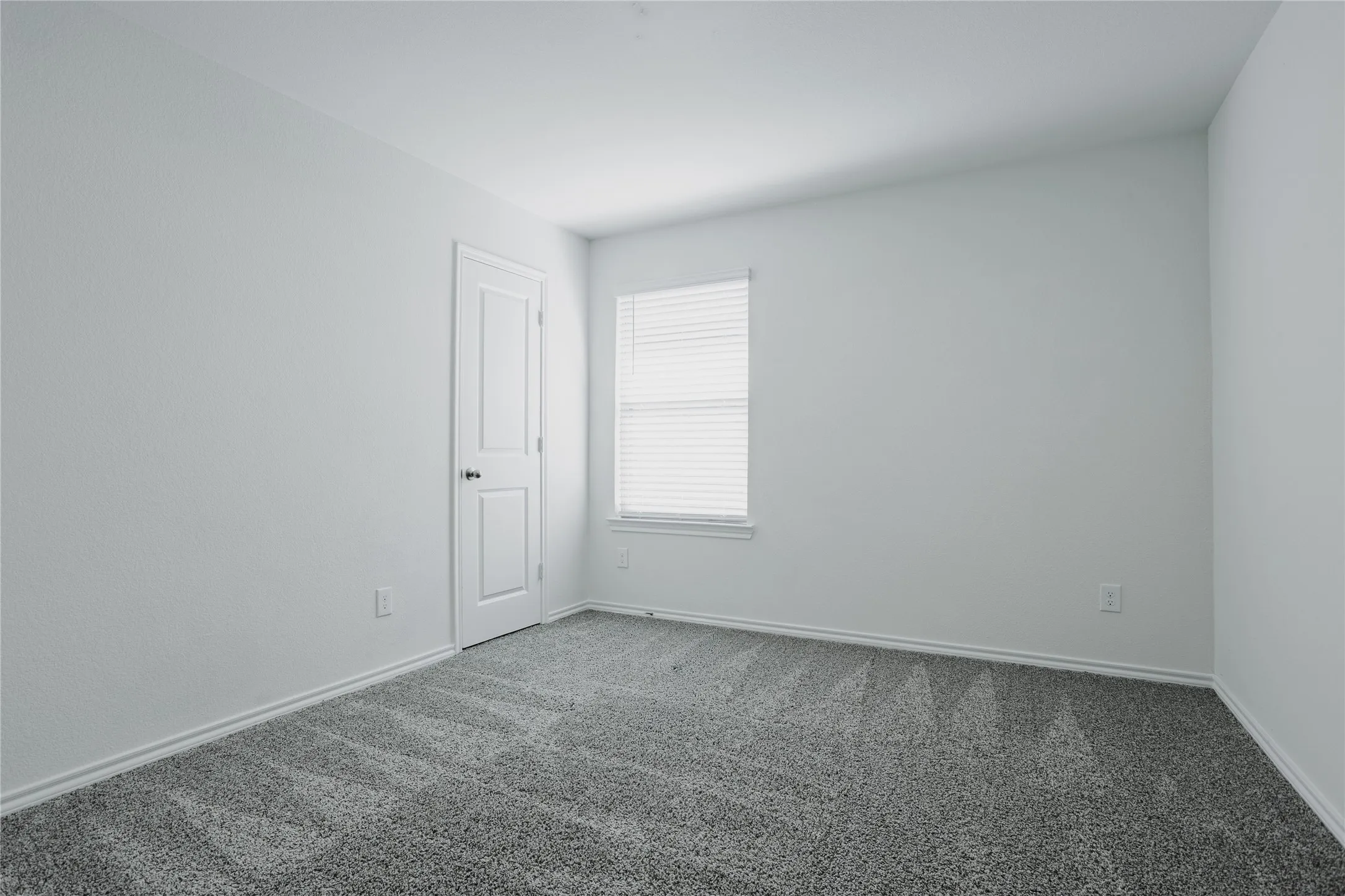 Neutral-toned room featuring a single window with blinds, light gray textured walls, and a gray patterned carpet