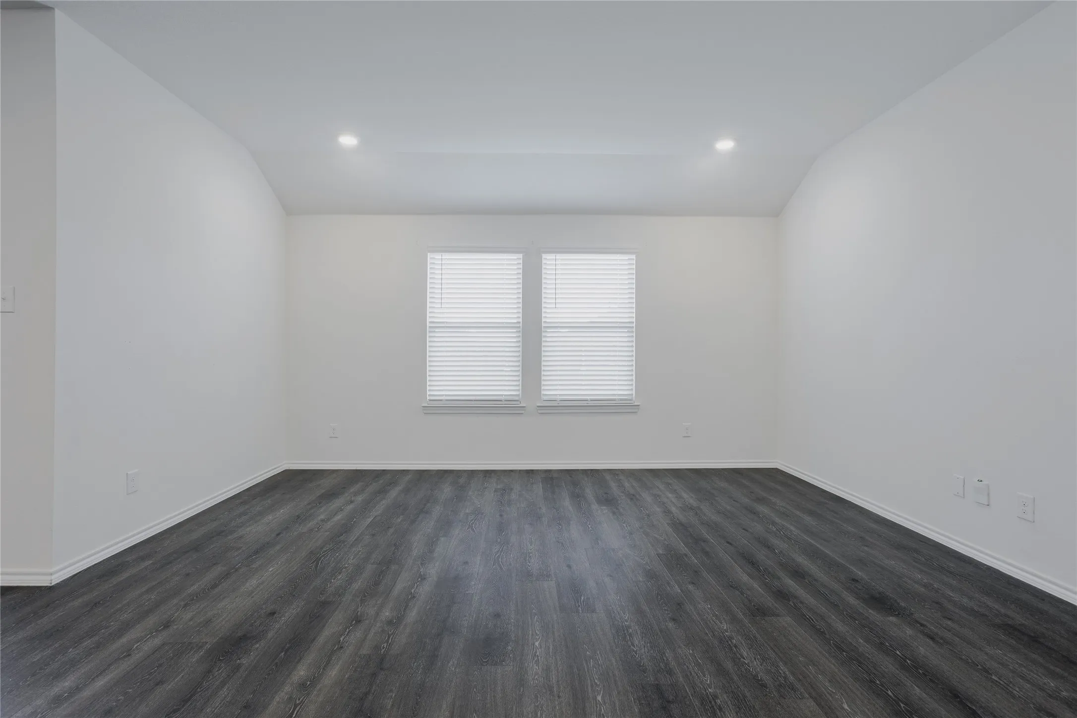 Spacious room featuring dark wood-finish flooring, white walls, and a window with blinds