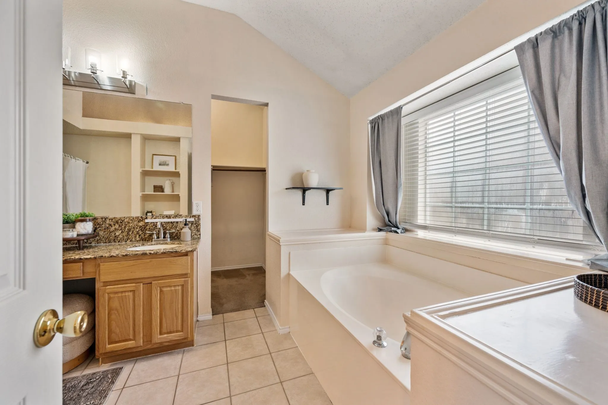 Spacious bathroom featuring a large built-in soaking tub, a single vanity with a granite-style countertop, natural wood cabinetry, and tiled flooring