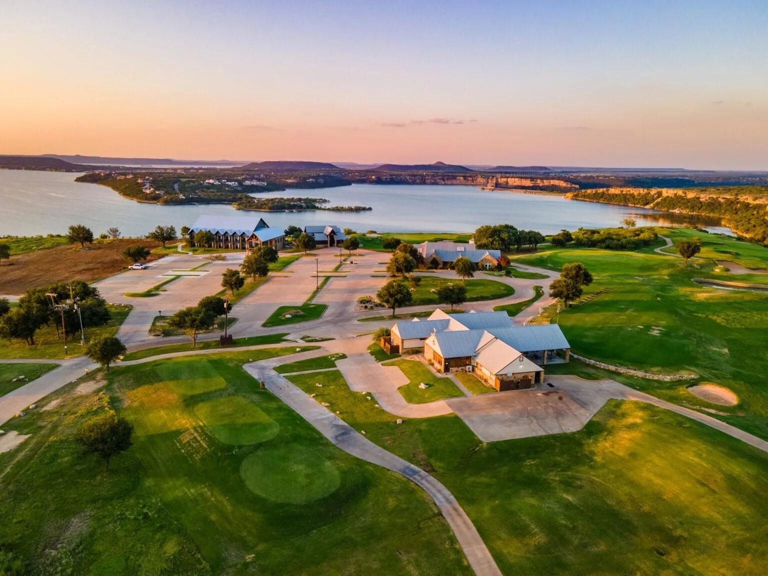 Expansive golf course property featuring multiple buildings with stone facades and metal roofs, extensive paved parking, and manicured green spaces