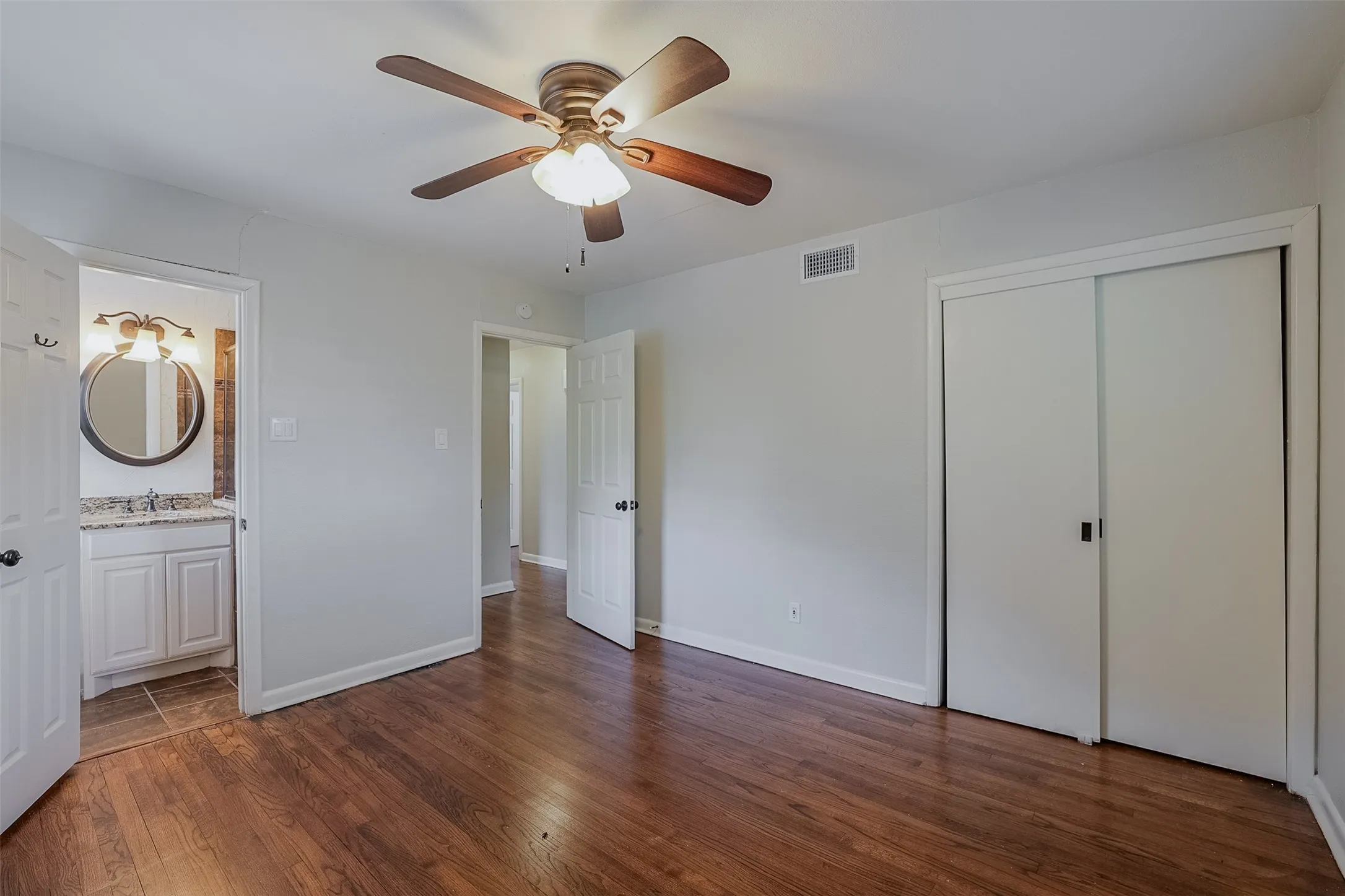 Wood-finish flooring, ceiling fan, white sliding closet doors, built-in vanity with granite-style countertop, and circular mirror