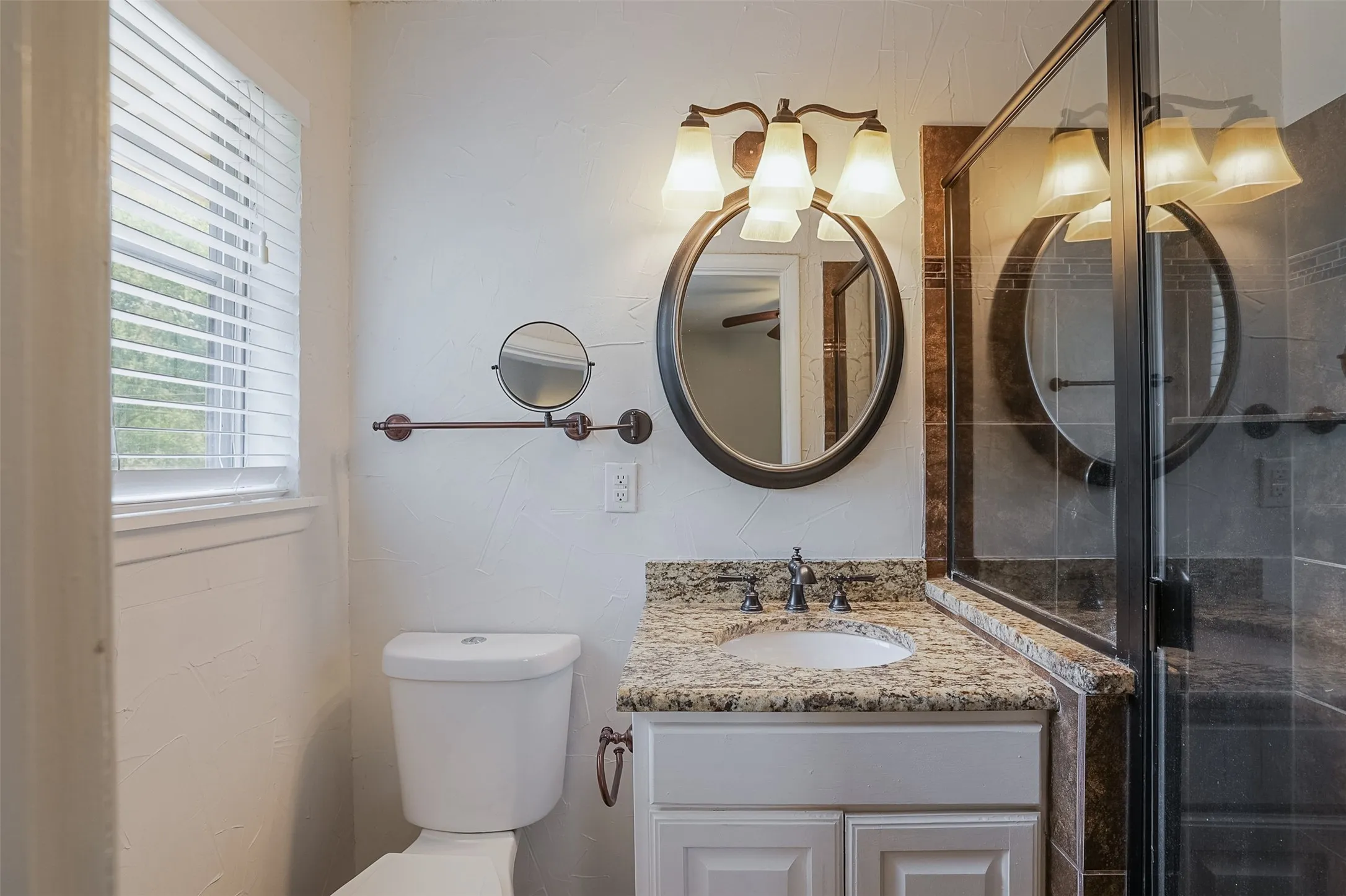 Vanity with granite countertop and integrated sink, framed mirror with overhead vanity lighting, glass-enclosed shower, and window with blinds