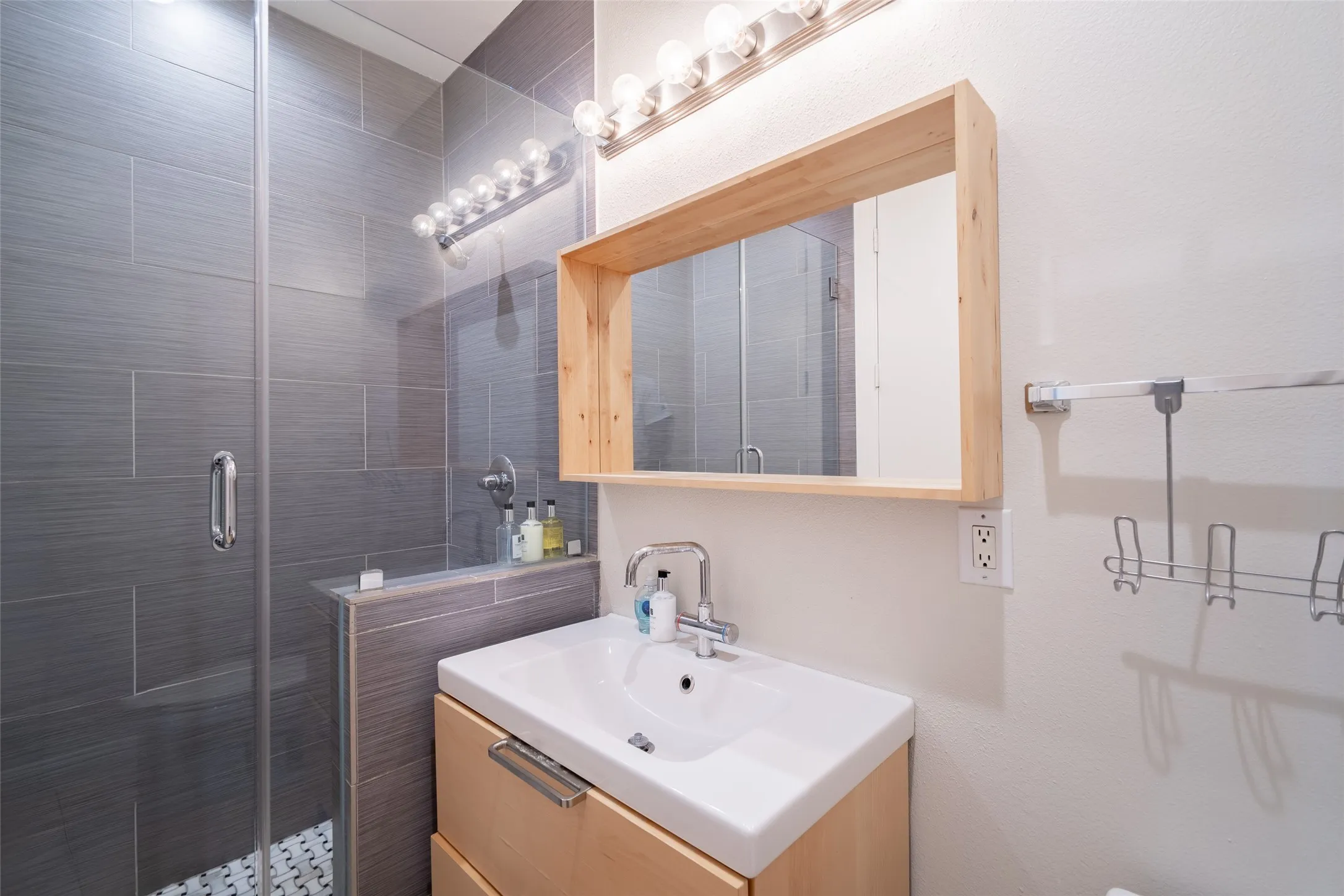 Bathroom featuring a glass-enclosed shower with gray tile, a light wood-finish vanity with an integrated sink, a rectangular mirror framed in light wood, and chrome fixtures