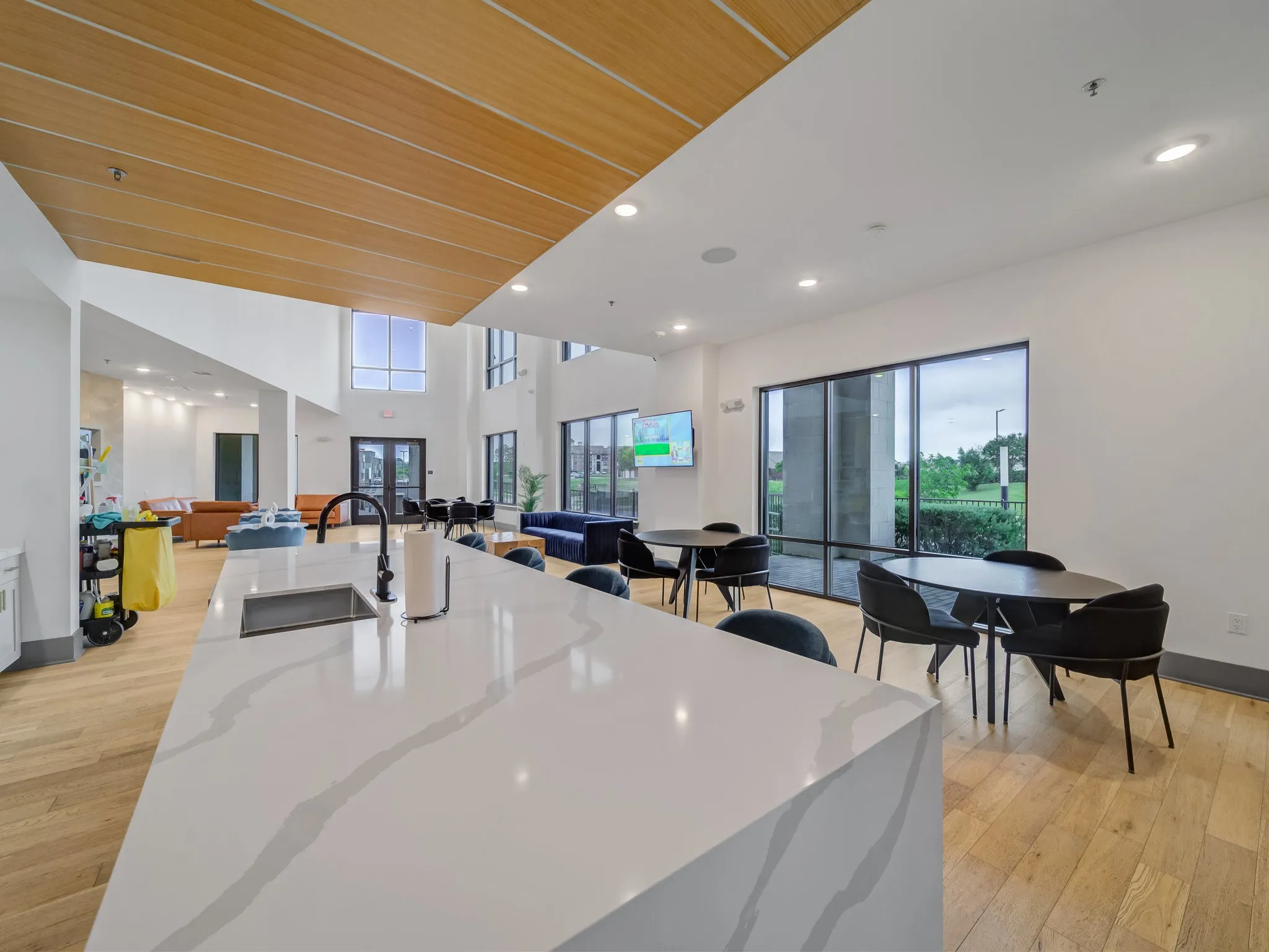 Spacious communal area featuring a large central island with an undermount sink and matte black faucet, wood-finish flooring, full-height windows, a wood-paneled ceiling accent, and recessed lighting