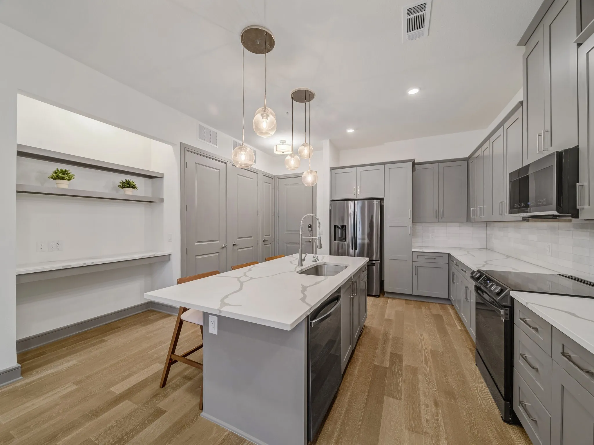 Open-concept kitchen featuring an oversized island with pendant lighting, light gray cabinetry with dark hardware, stainless steel appliances, and wood-finish flooring
