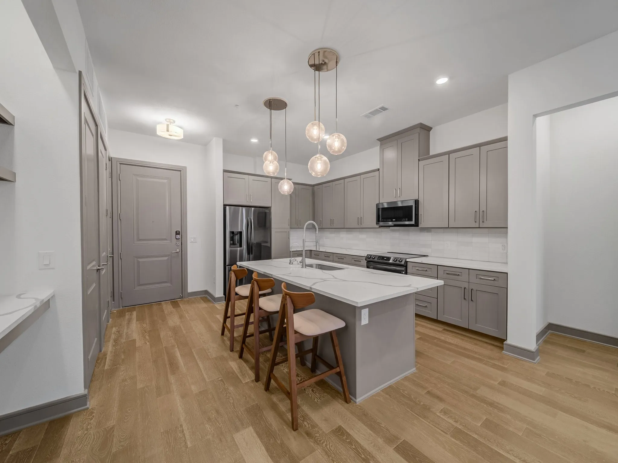 Contemporary kitchen featuring a large central island with seating, pendant lighting, light gray cabinetry, stainless steel appliances, and wood-finish flooring