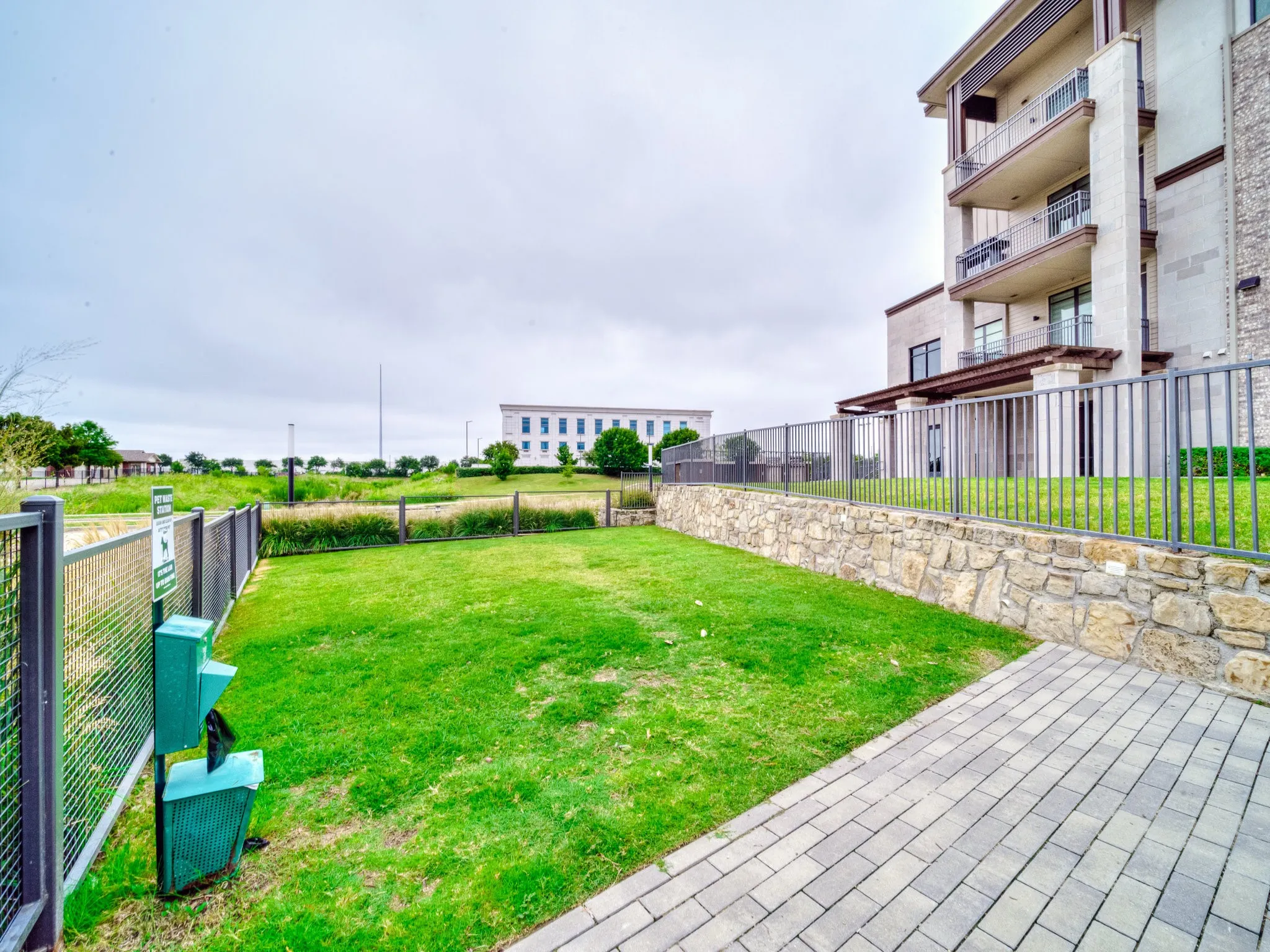 Grassed outdoor area featuring a paved pathway, stone retaining wall, and metal fencing