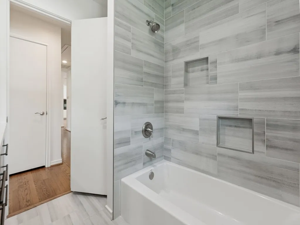 Bathtub and shower combination featuring gray wood-finish tiled walls, two recessed shower niches, and a chrome shower fixture