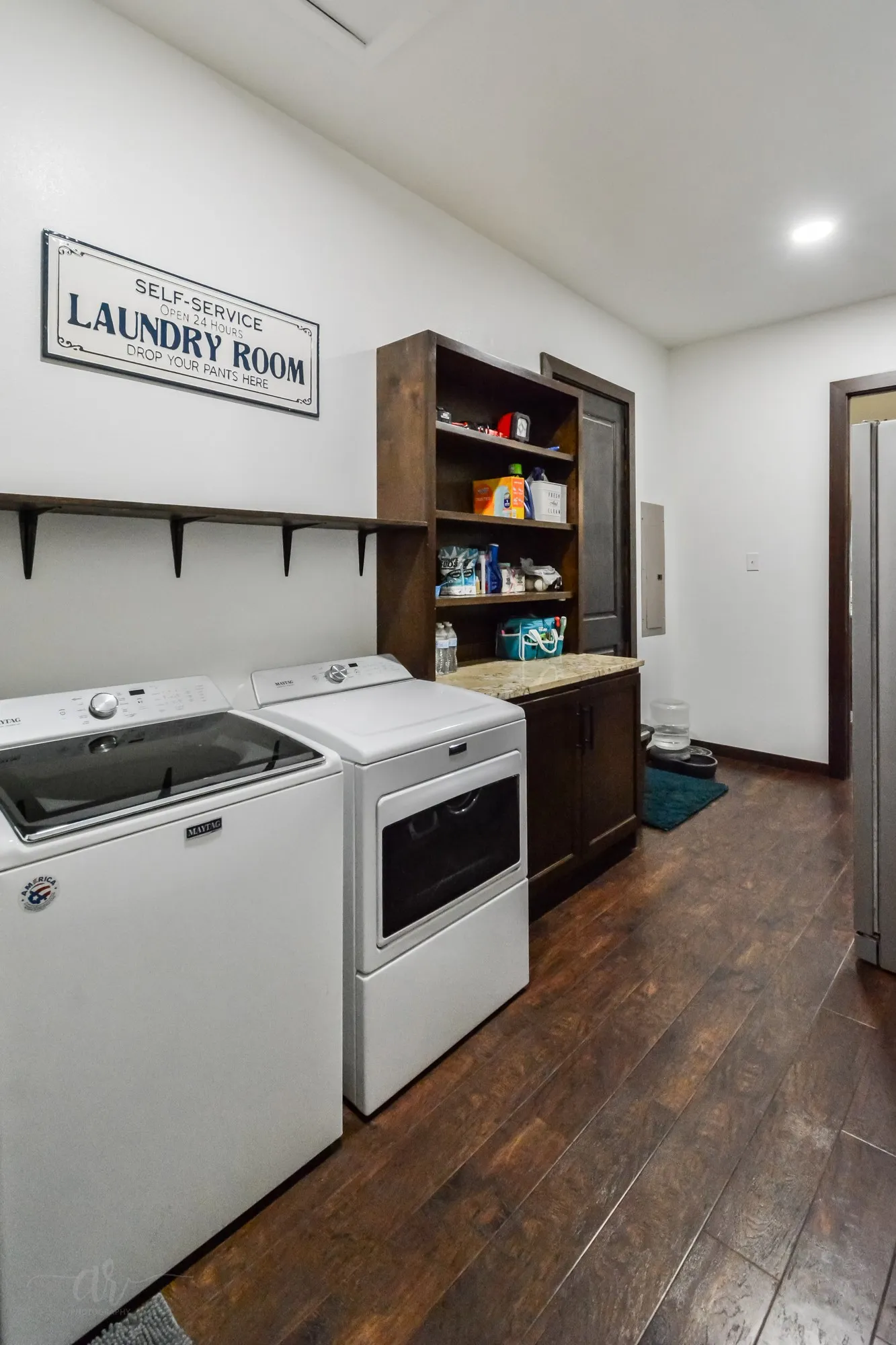 Dedicated laundry area featuring wood-finish flooring, a built-in shelving unit, dark wood cabinetry, and a countertop surface