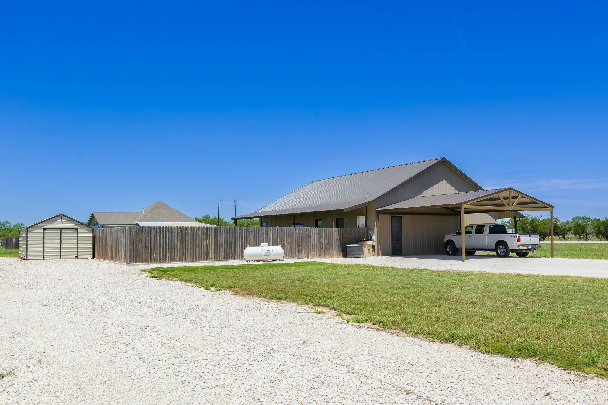 Property featuring a metal roof, attached carport, privacy fencing, storage shed, and gravel driveway