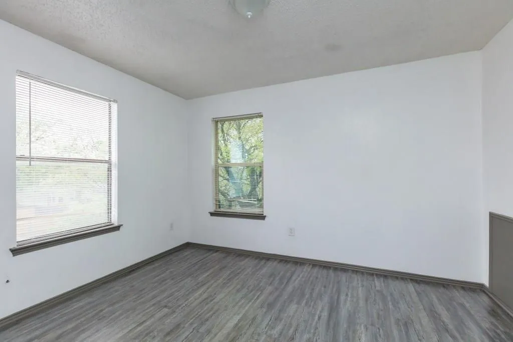 Spacious room featuring two windows with horizontal blinds, wood-finish flooring, white walls, and a ceiling-mounted light fixture