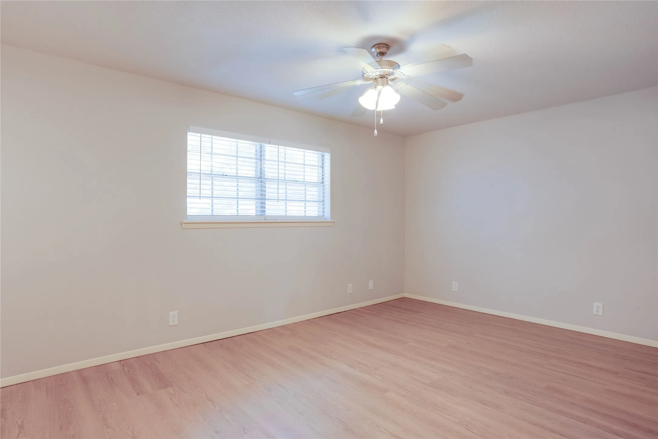 Spacious room featuring light-colored walls, wood-finish flooring, a window with horizontal blinds, and a ceiling fan with integrated lighting
