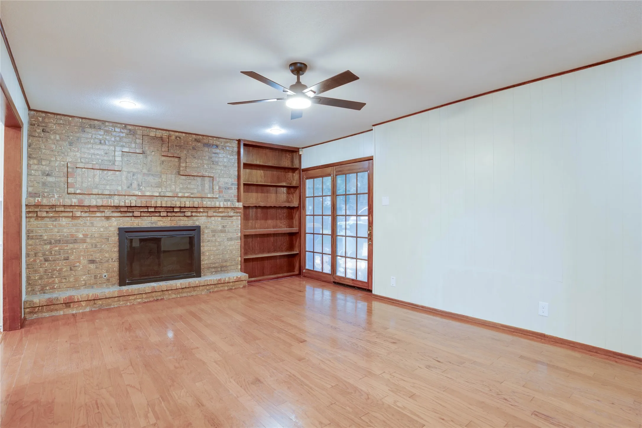 Spacious room featuring wood-finish flooring, a brick fireplace, built-in shelving, glass-paned double doors, and a ceiling fan with integrated lighting