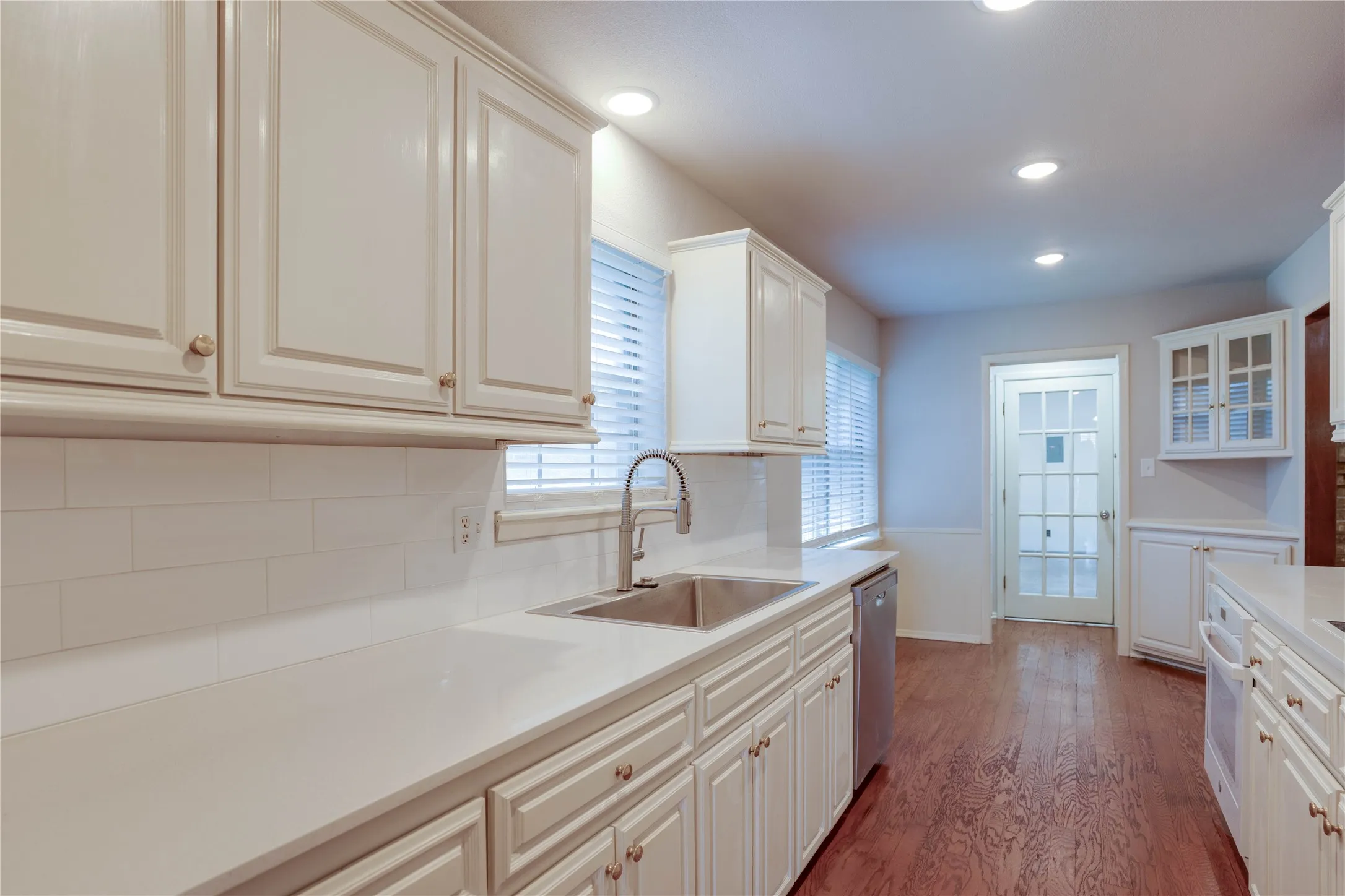 White cabinetry with gold-tone hardware, white countertops, subway tile backsplash, stainless steel double basin sink, and wood-finish flooring