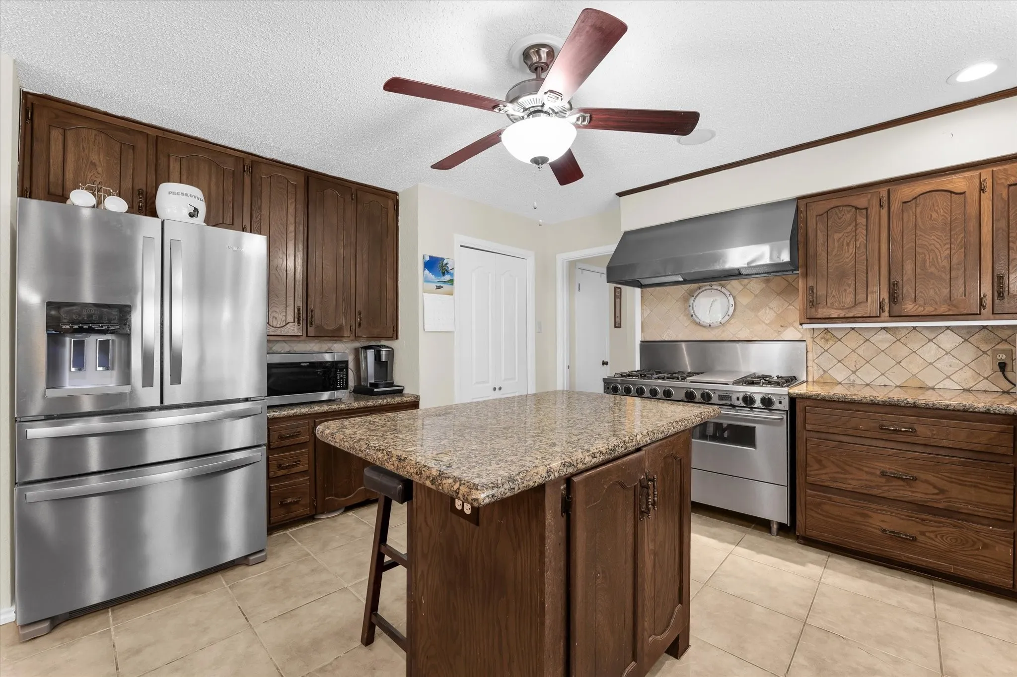 Kitchen featuring wood-finish cabinetry, granite-style countertops, stainless steel appliances, a tiled backsplash, and a central island