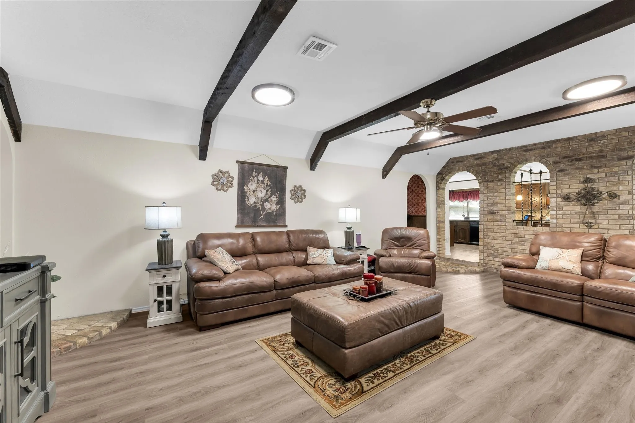 Spacious living area featuring exposed ceiling beams, wood-finish flooring, a ceiling fan, and an accent brick wall with arched openings