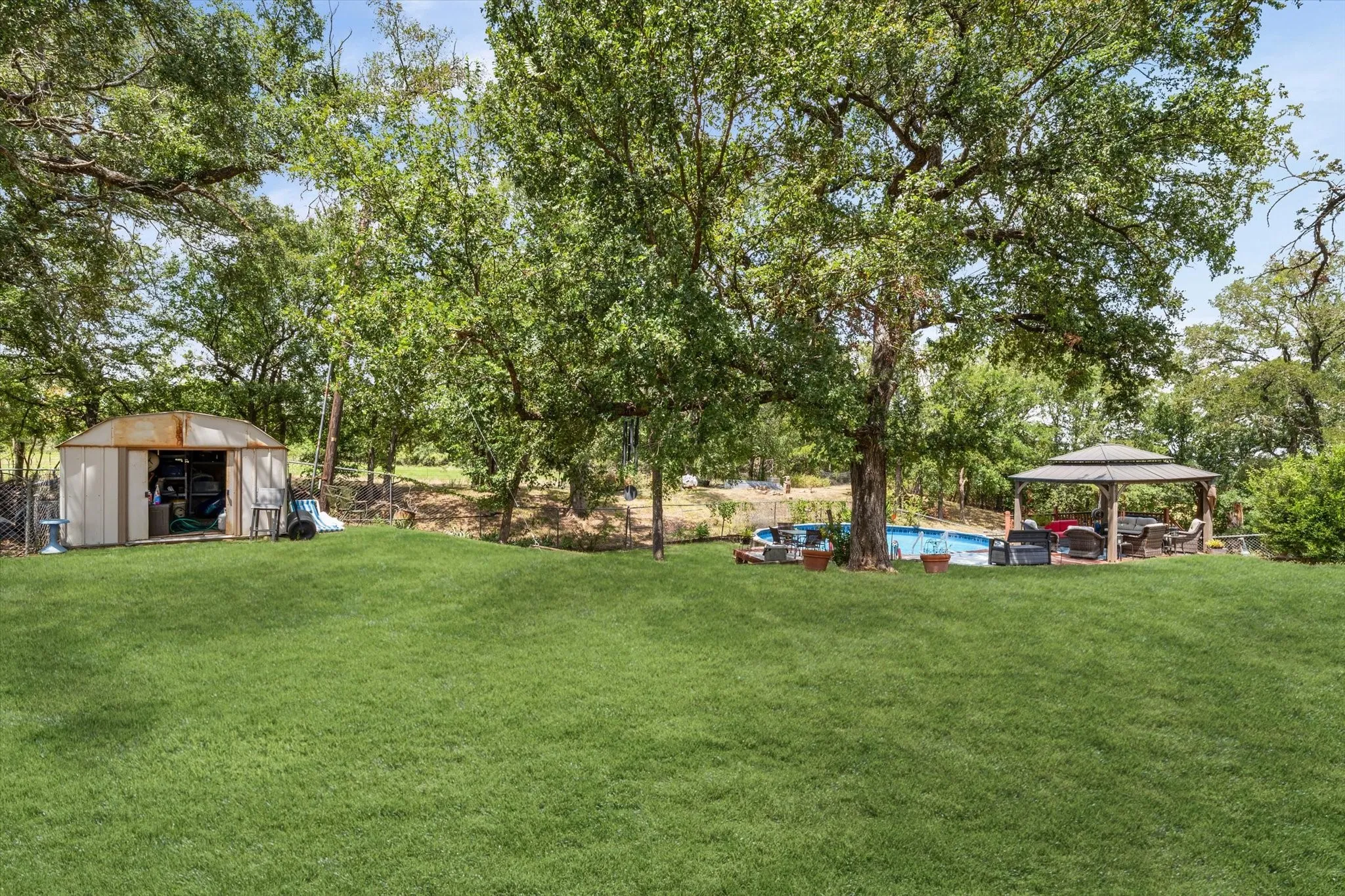 Expansive outdoor area featuring a swimming pool, a covered gazebo, mature trees, and a storage shed