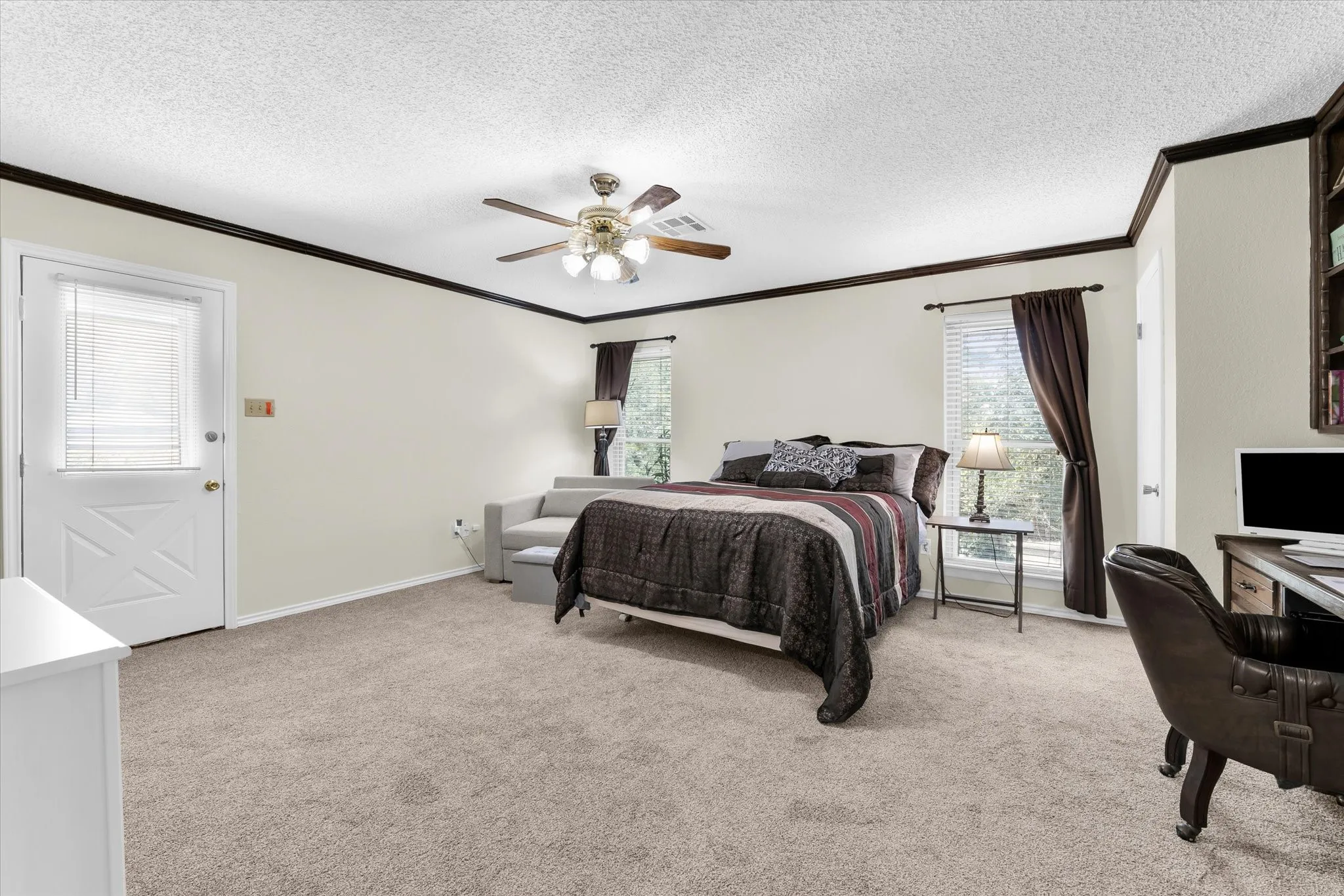 Carpeted room featuring a ceiling fan with light kit, dark wood trim, built-in shelving, two windows with blinds, and a white exterior door with window insert