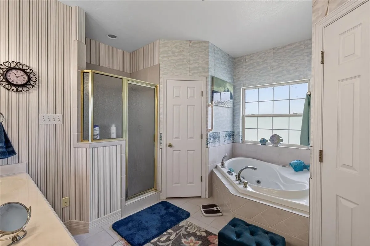 Bathroom featuring an integrated corner jetted tub, separate shower enclosure with gold-tone framing, tiled tub surround, and a window
