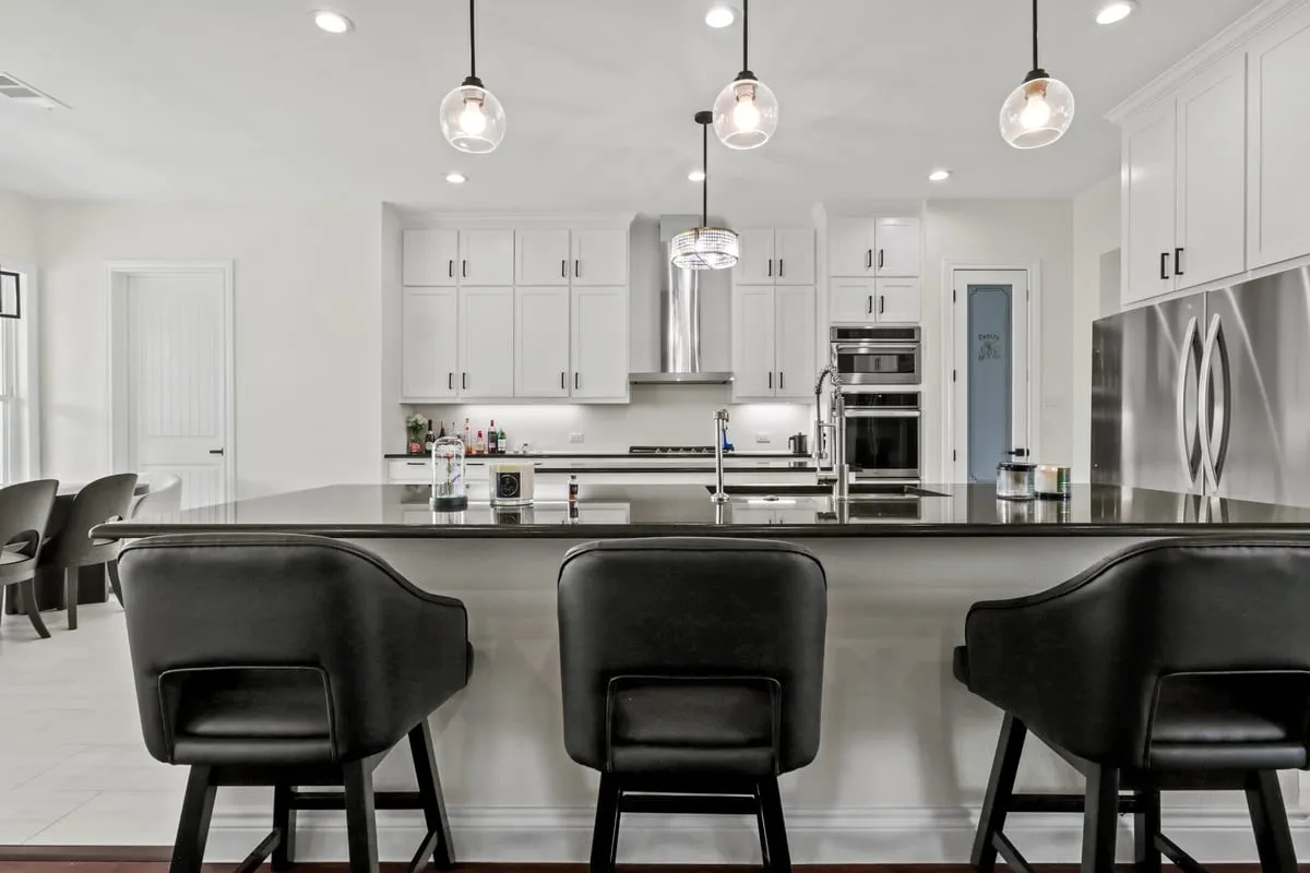 Contemporary kitchen featuring a large center island with waterfall-edge countertop, three pendant lights with clear glass shades, custom white cabinetry with chrome hardware, stainless steel appliances including a double wall oven configuration, and a professional-grade range hood
