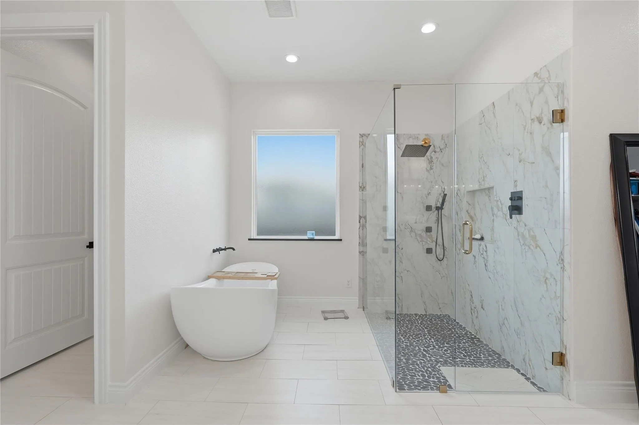 Freestanding soaking tub, large window, glass-enclosed shower with marble-look wall tiles, pebble tile shower floor, and multiple shower heads