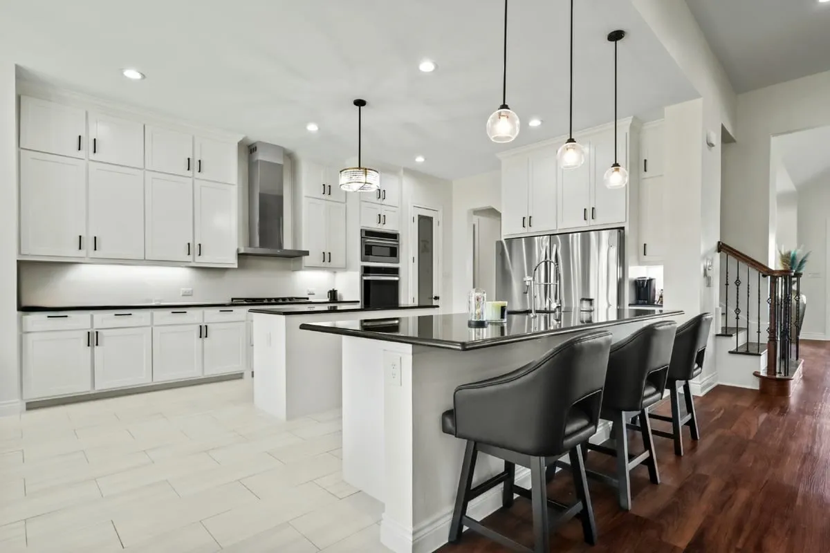 Modern kitchen featuring white shaker cabinetry with black hardware, stainless steel appliances including a built-in double wall oven, quartz countertops, a large center island with seating for multiple guests, pendant lighting, and hardwood flooring throughout the open-concept space
