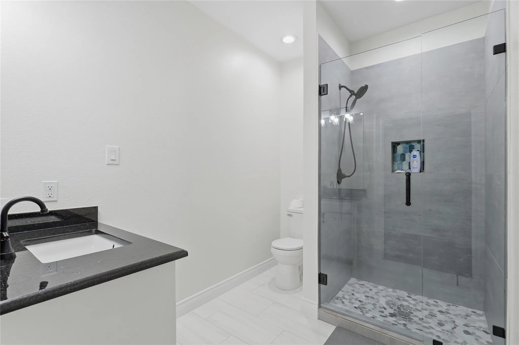 Modern bathroom featuring a glass-enclosed shower with pebble-tile flooring, a built-in shower niche, and a vanity with a dark countertop and undermount sink
