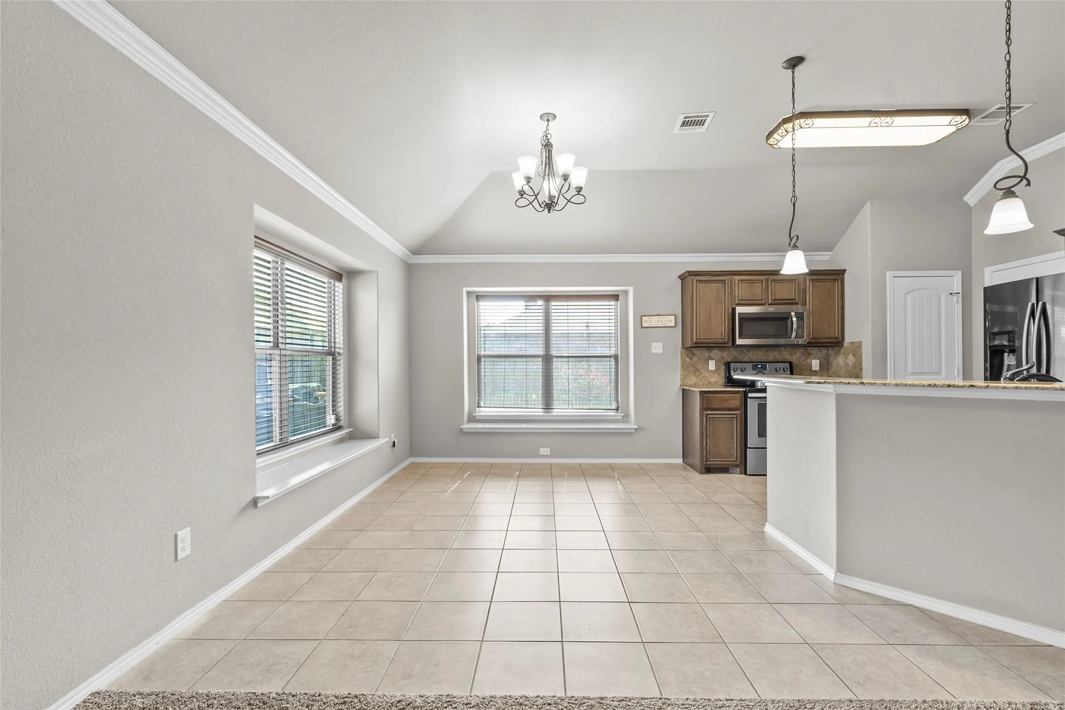Open concept living space featuring tile flooring throughout, vaulted ceiling, crown molding, and two large windows with blinds