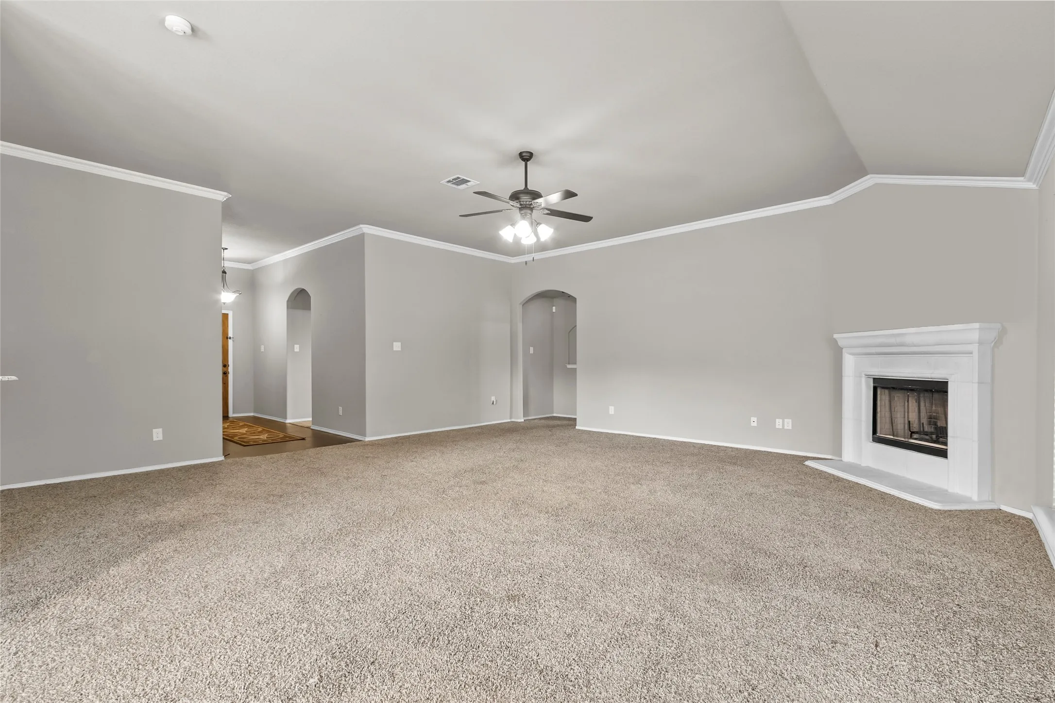 Spacious carpeted room featuring a white mantel fireplace, a ceiling fan with integrated lighting, and crown molding
