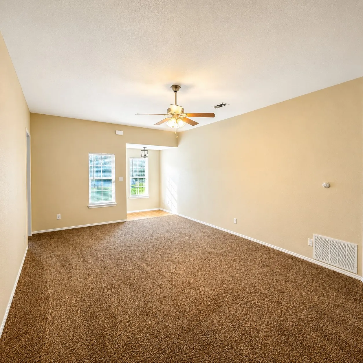 Spacious room with plush brown carpeting, light tan wall color, and a ceiling fan with wood-finish blades