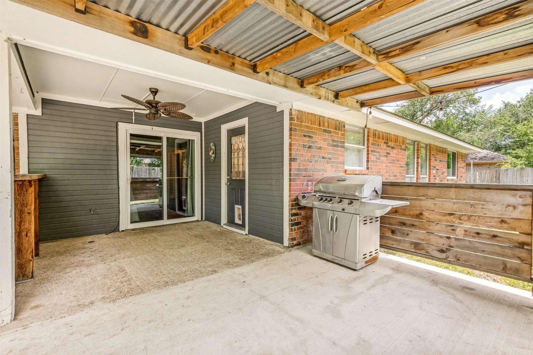 Covered concrete patio featuring a corrugated metal roof supported by exposed wood beams, a ceiling fan, and sliding glass doors