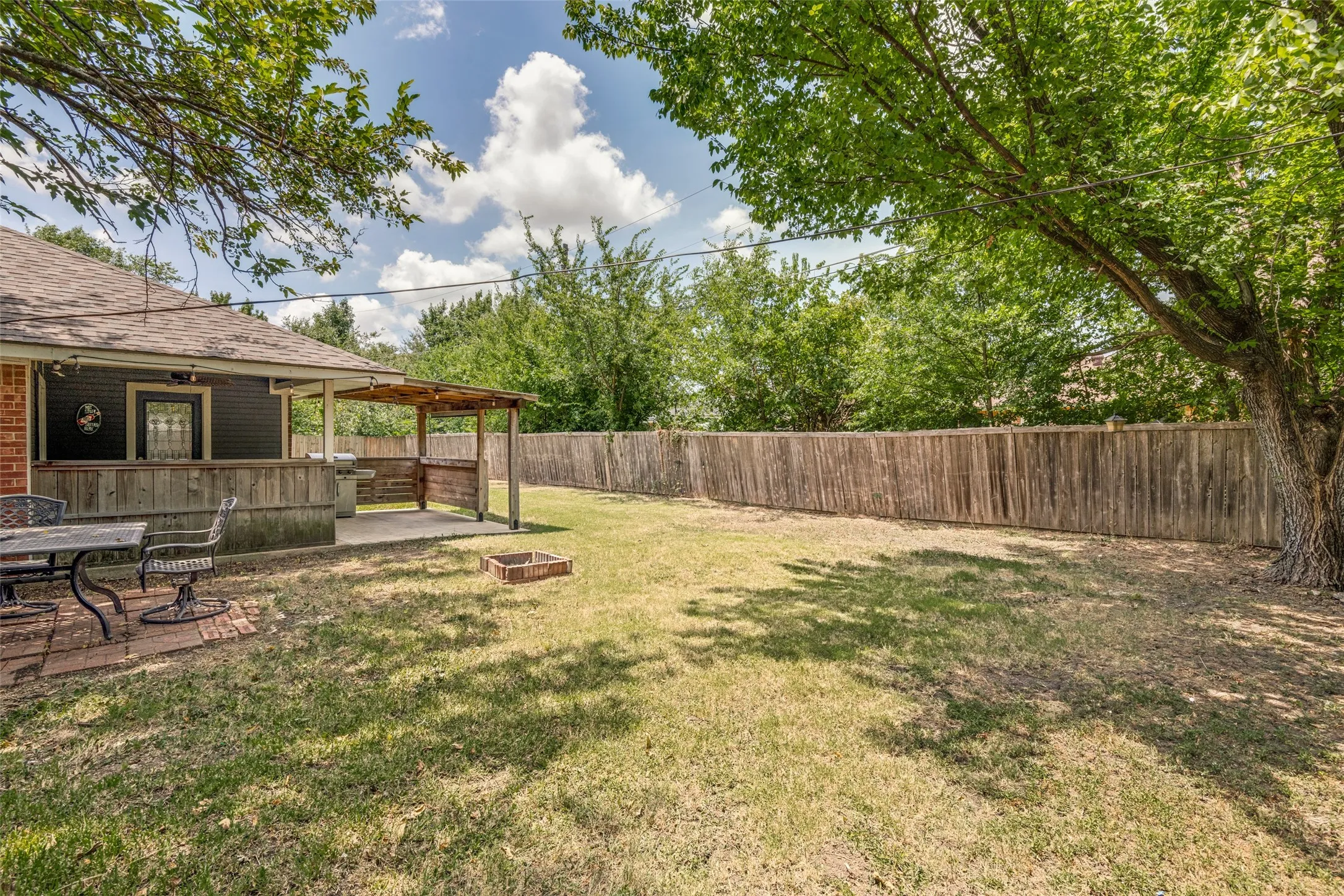 Expansive backyard featuring a covered patio with built-in grill, a separate brick paver patio, mature trees providing natural shade, and a full privacy fence