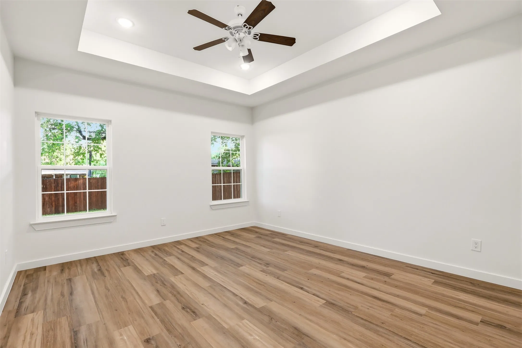 Spacious room featuring wood-finish flooring, two large windows, a tray ceiling with recessed lighting, and a ceiling fan