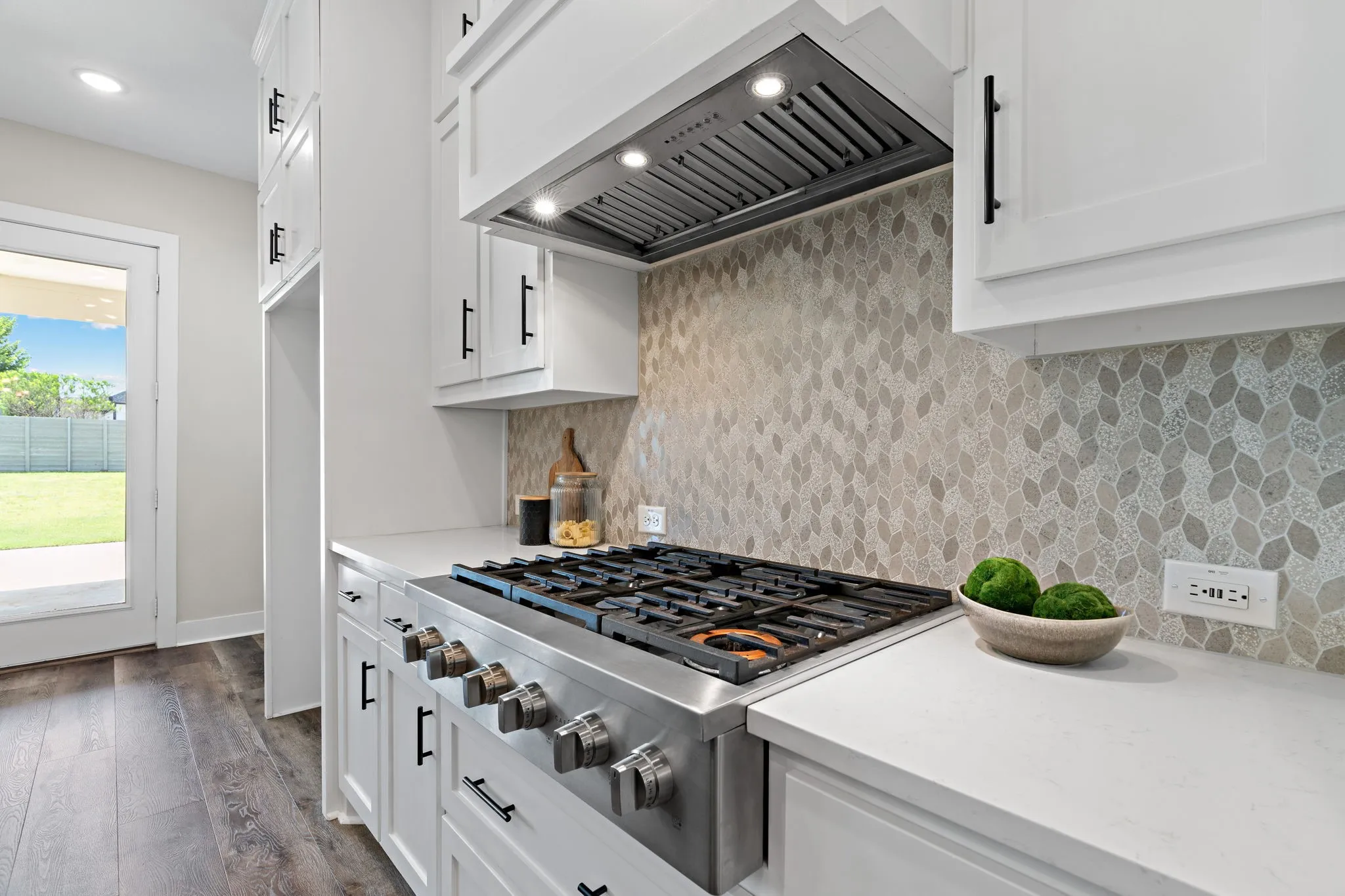 Stainless steel gas range with coordinating ventilation hood, white cabinetry with matte black hardware, mosaic tile backsplash, light-toned countertops, and wood-finish flooring