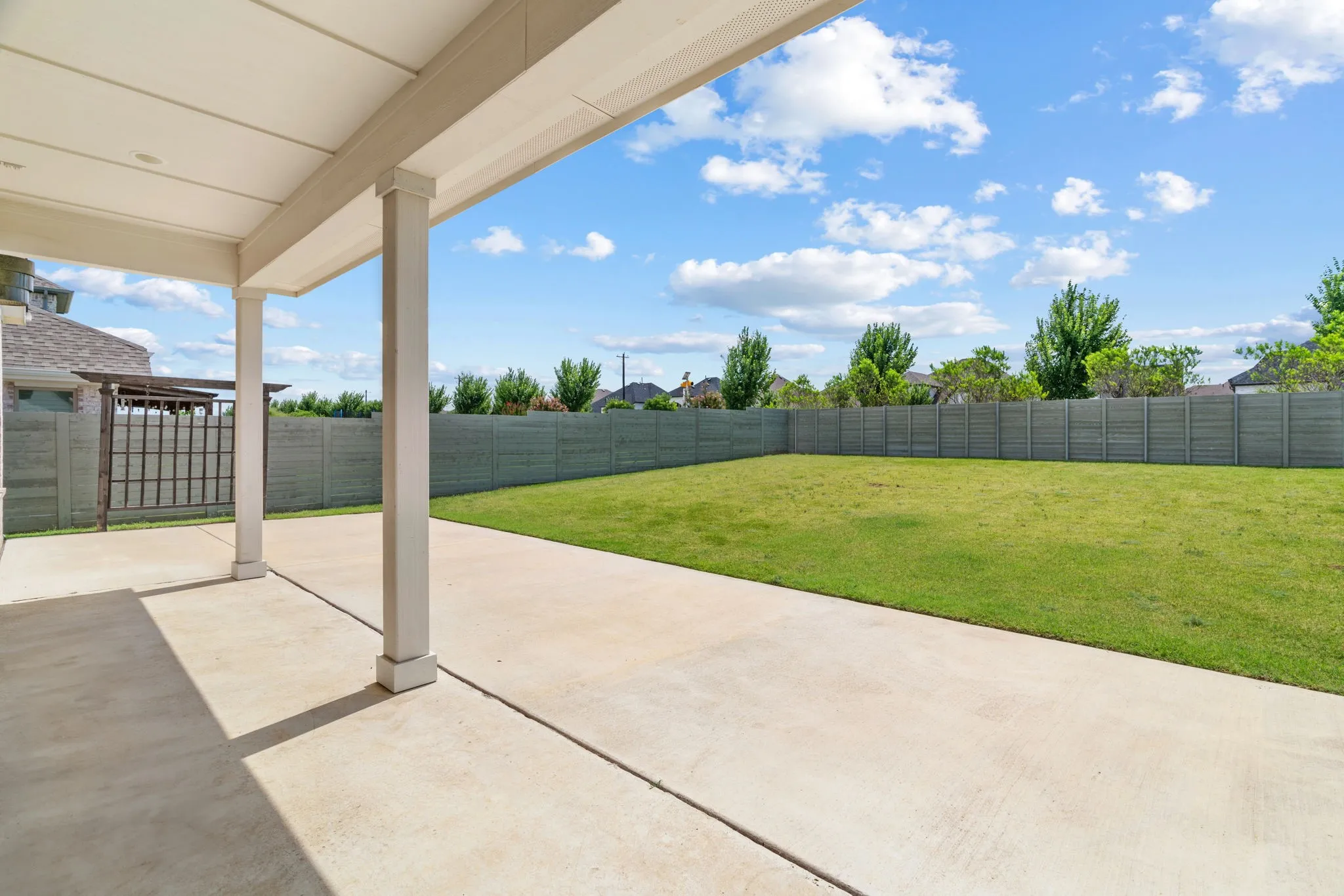 Expansive concrete patio with a covered outdoor living area and a large grassy backyard featuring mature trees and a privacy fence