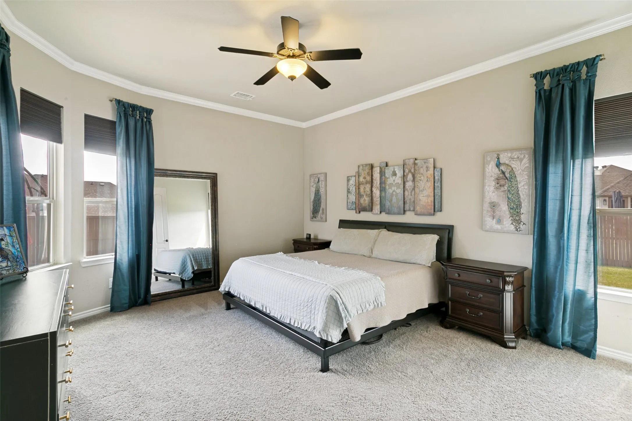 Spacious Primary bedroom showcasing a coffered ceiling with integrated ceiling fan, abundant natural light from dual windows dressed in teal curtains, neutral wall finishes, and neutral carpet flooring