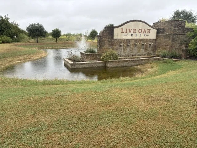 Stone entry monument featuring "Live Oak Creek" lettering, water feature, adjacent pond, and established trees