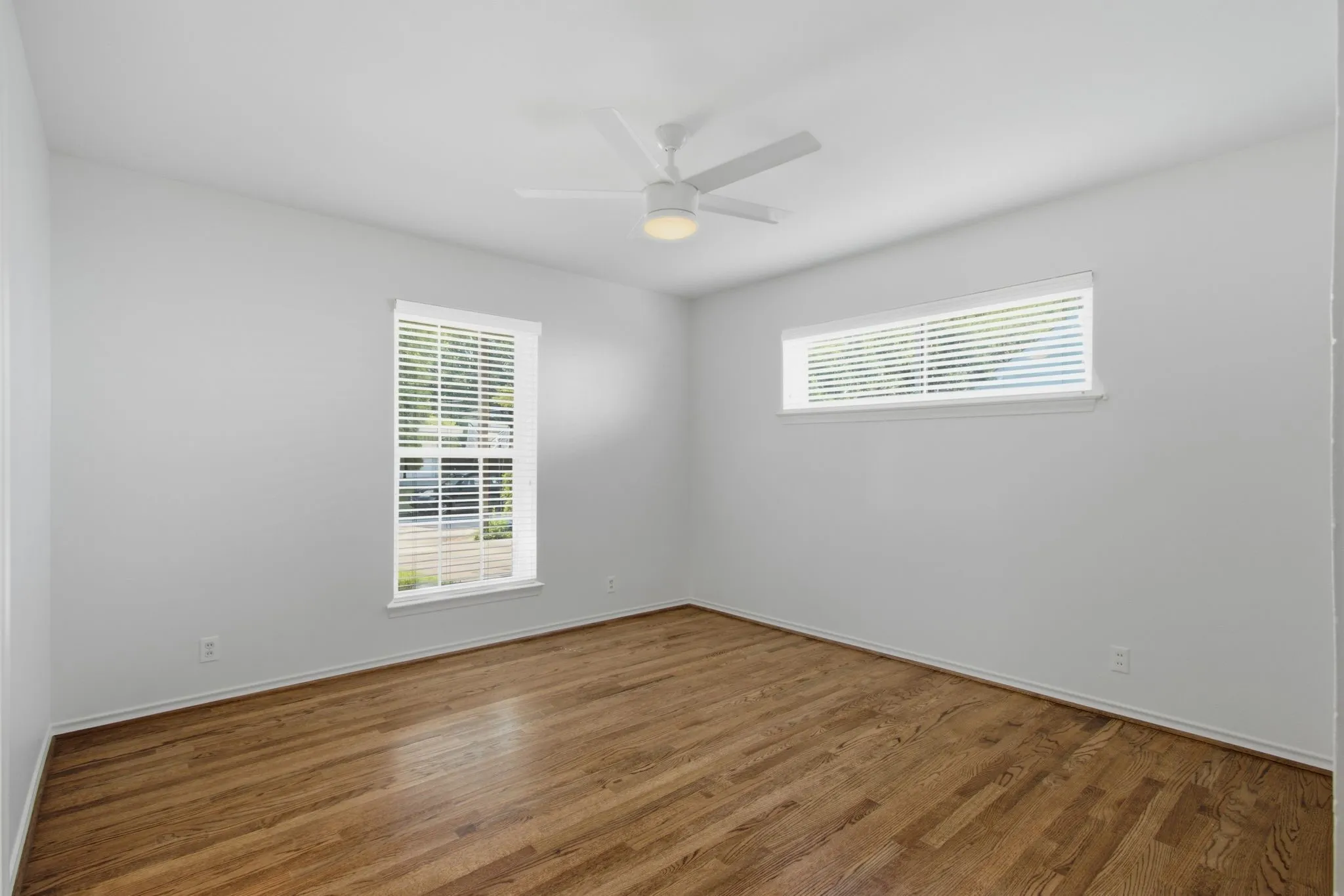 Bright room with wood-finish flooring, a contemporary ceiling fan with integrated lighting, a rectangular window with blinds, and an additional horizontal window, all set against neutral wall tones