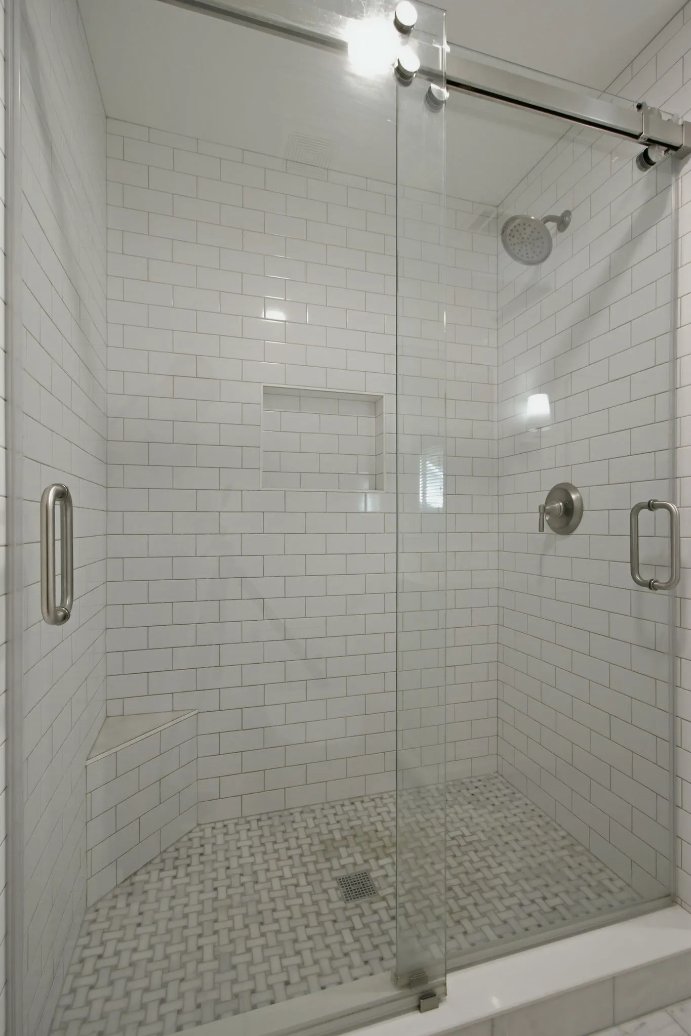 Walk-in shower featuring a frameless glass enclosure with brushed nickel hardware, white subway tile walls, a built-in shower niche, and a basketweave tile floor
