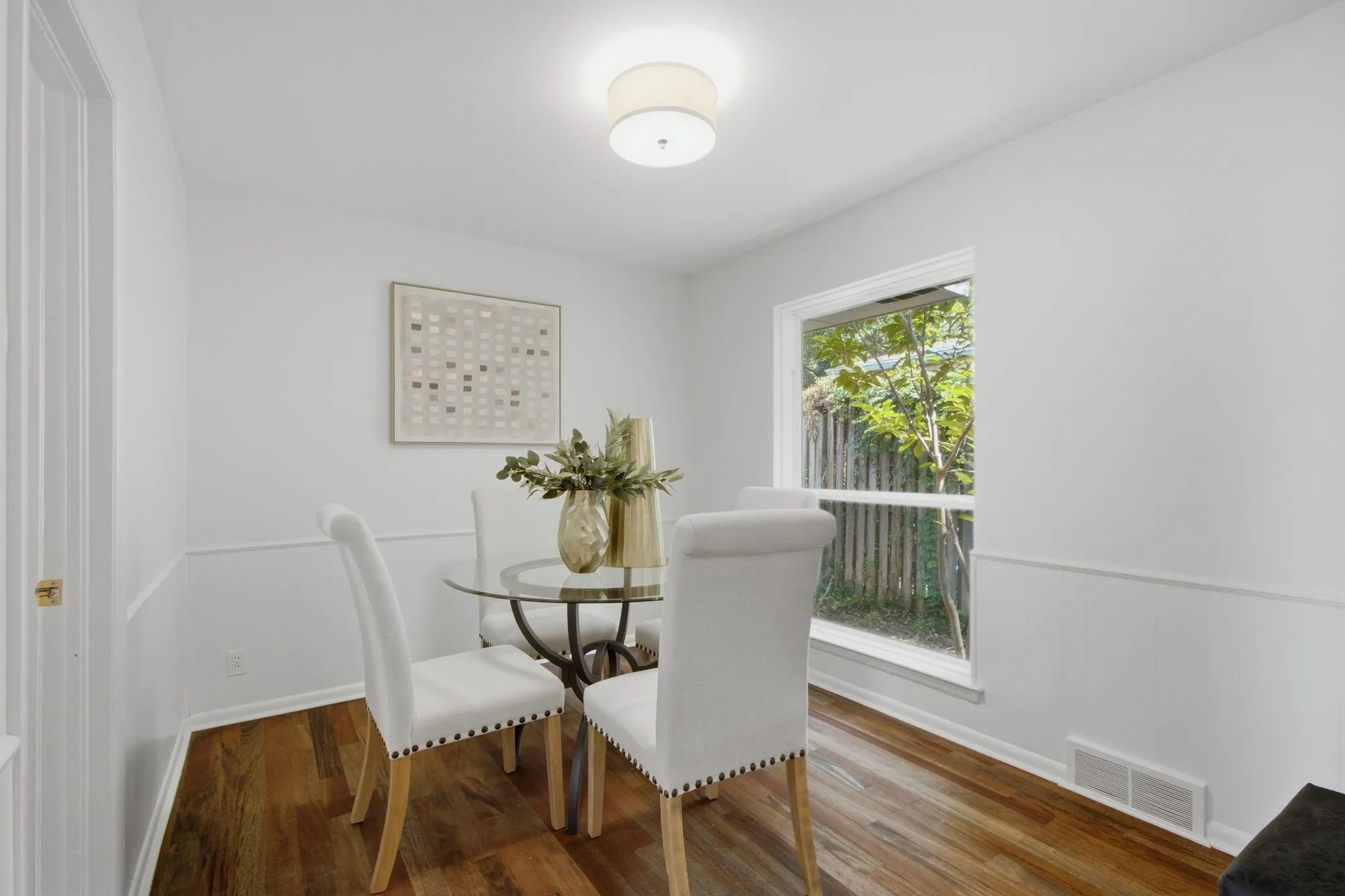 Dining area featuring wood-finish flooring, white paneled wainscoting, and a window with white trim overlooking exterior foliage