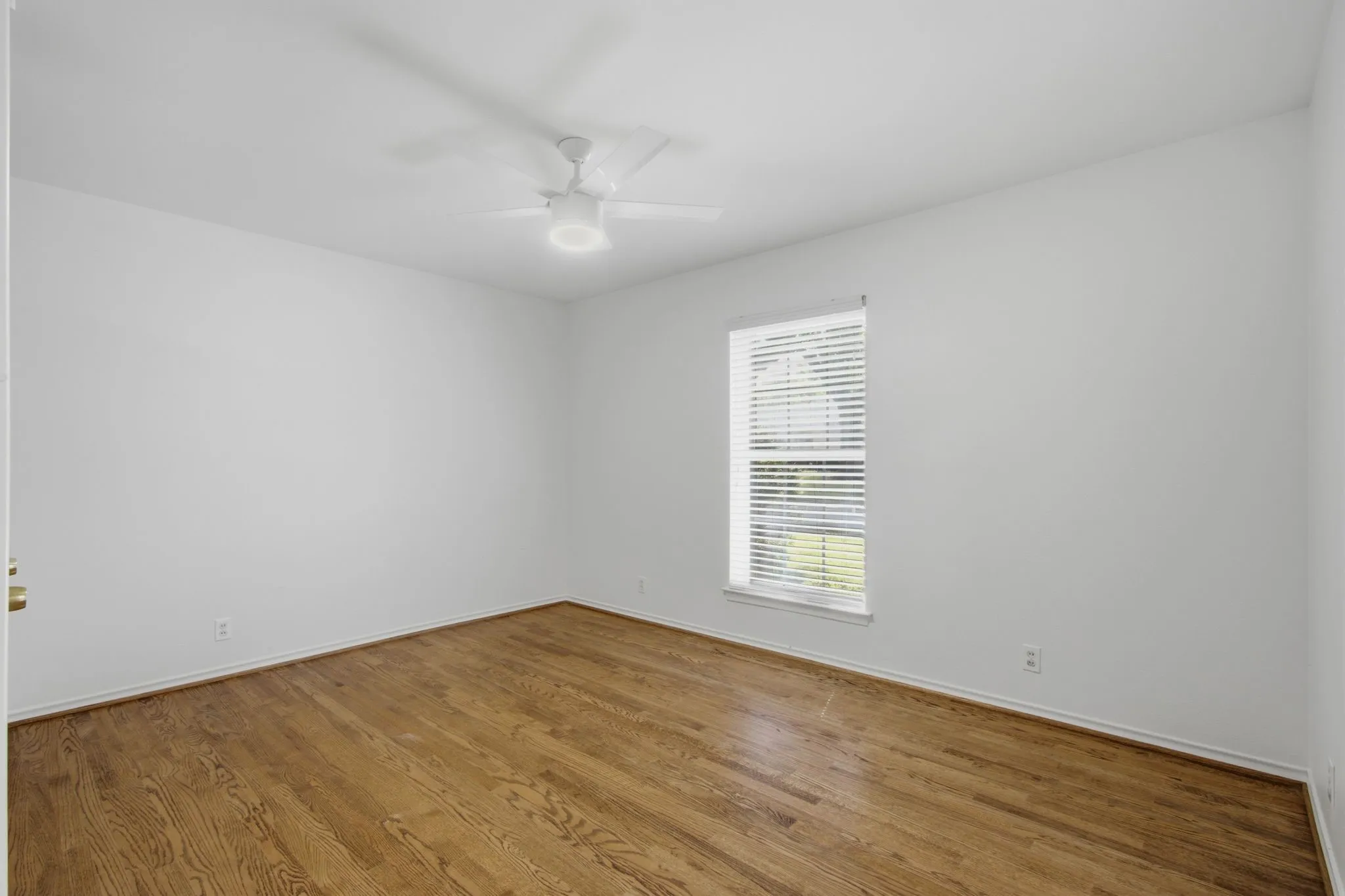 Spacious room featuring wood-finish flooring, crisp white walls, and a large window with horizontal blinds