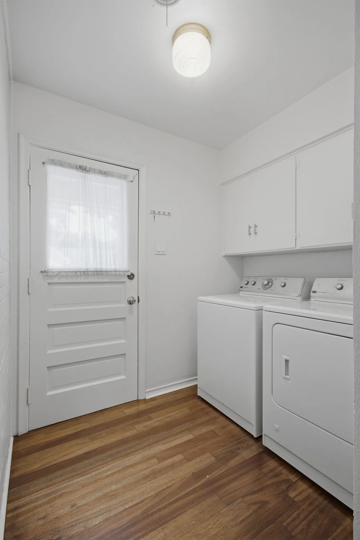 Utility room featuring wood-finish flooring, overhead cabinetry, and a solid panel door with a sheer curtain