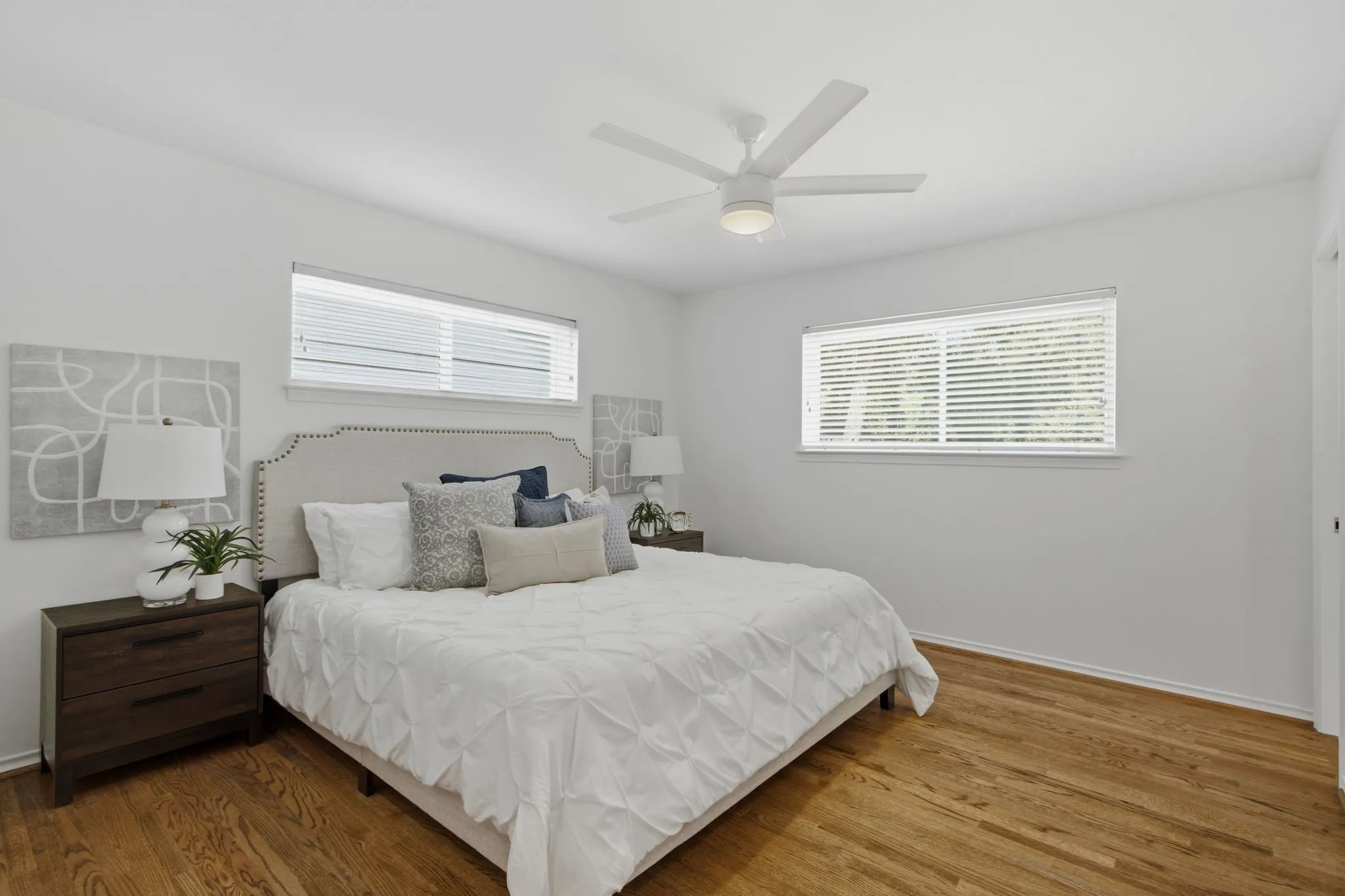 Bright interior featuring wood-finish flooring, white walls, two horizontal windows with blinds, and a white ceiling fan with integrated lighting