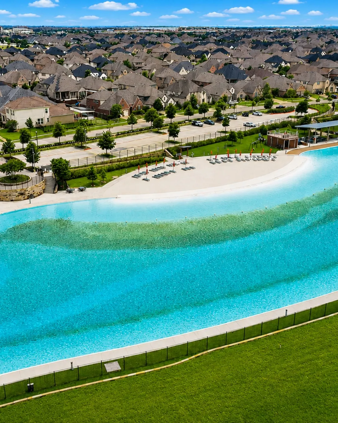 Expansive turquoise lagoon, sandy beach area, lush green lawn, adjacent residential properties, and paved parking areas