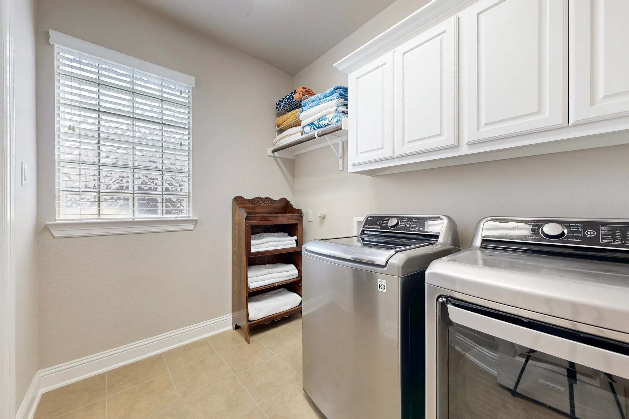 Dedicated laundry room featuring stainless steel front-load washer and dryer, white cabinetry with ample storage, tile flooring, and a large window providing natural light